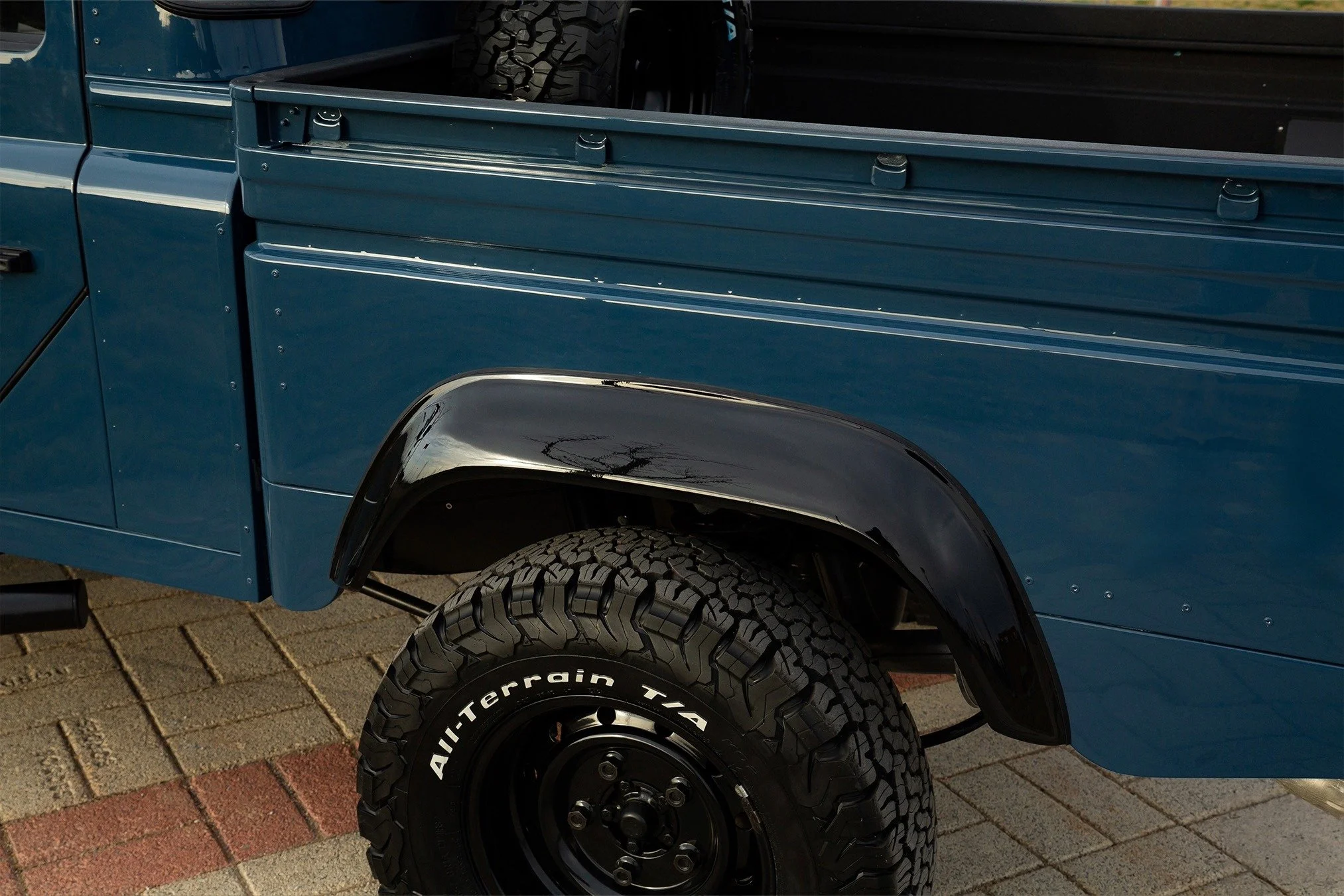 Close-up of the front wheel and fender of a blue off-road truck, with a black tire labeled 'All-Terrain T/A' and a dark fender flare.