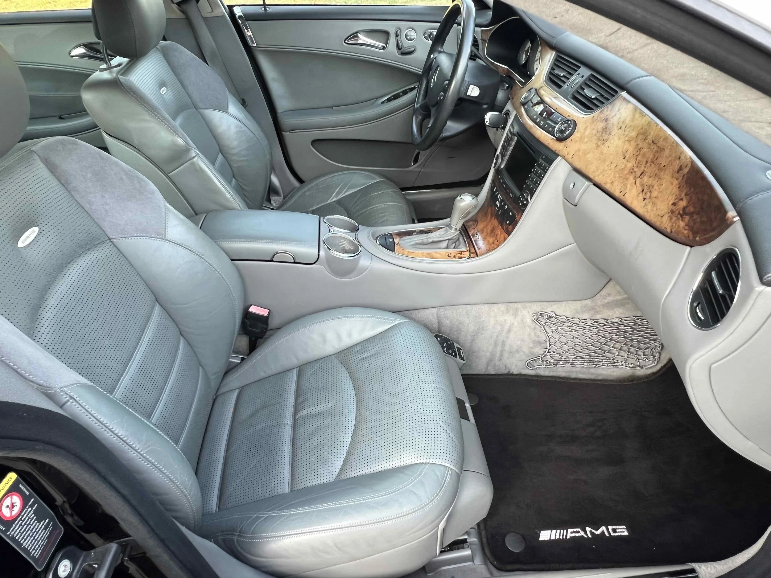 Interior of a Mercedes-Benz AMG car showing leather seats, wood trim dashboard, gear shift, center console with cup holders, and AMG branded floor mat.