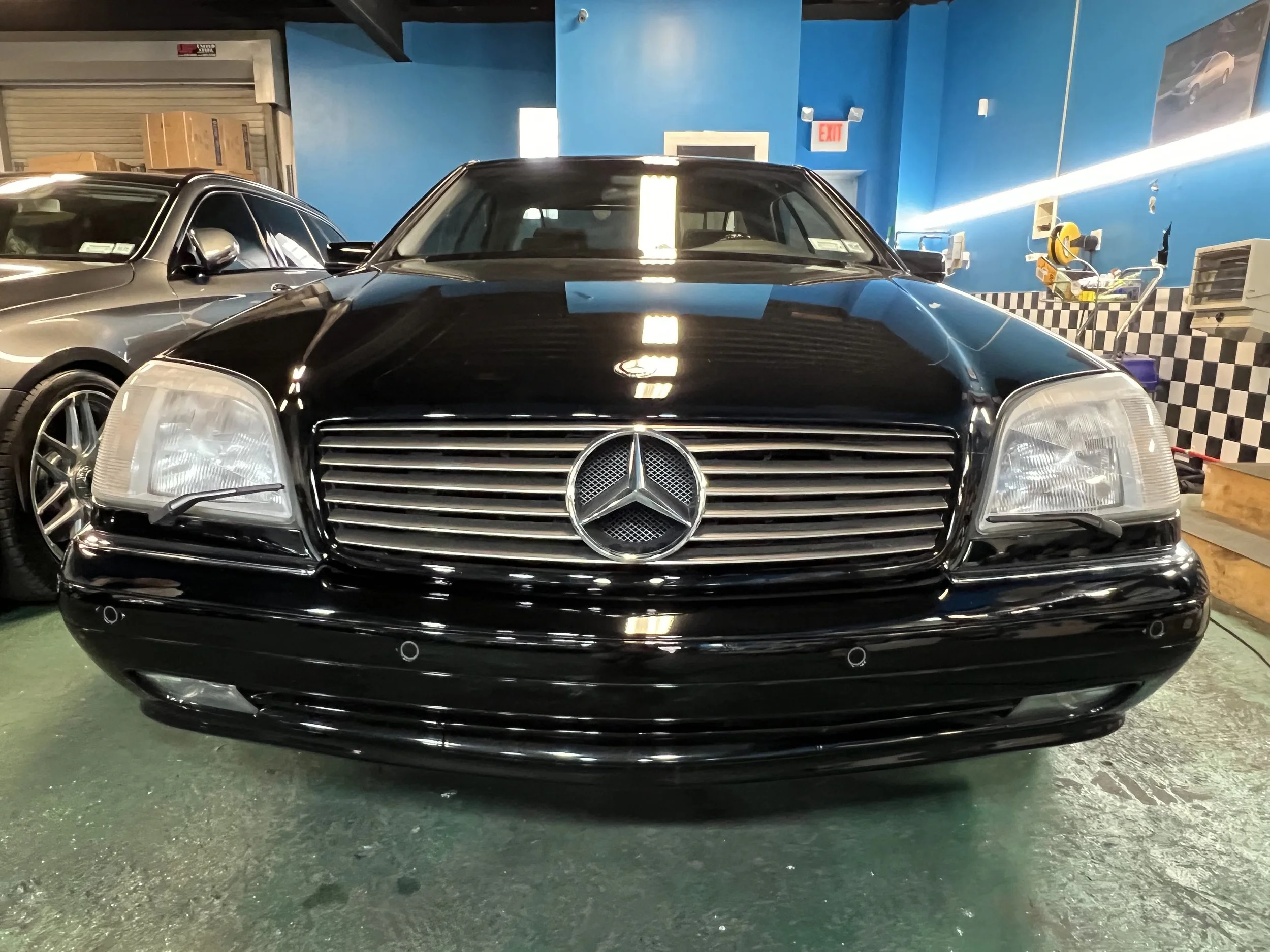 Black Mercedes-Benz car in a garage with other vehicles, blue walls, checkered black and white pattern, and various tools visible.