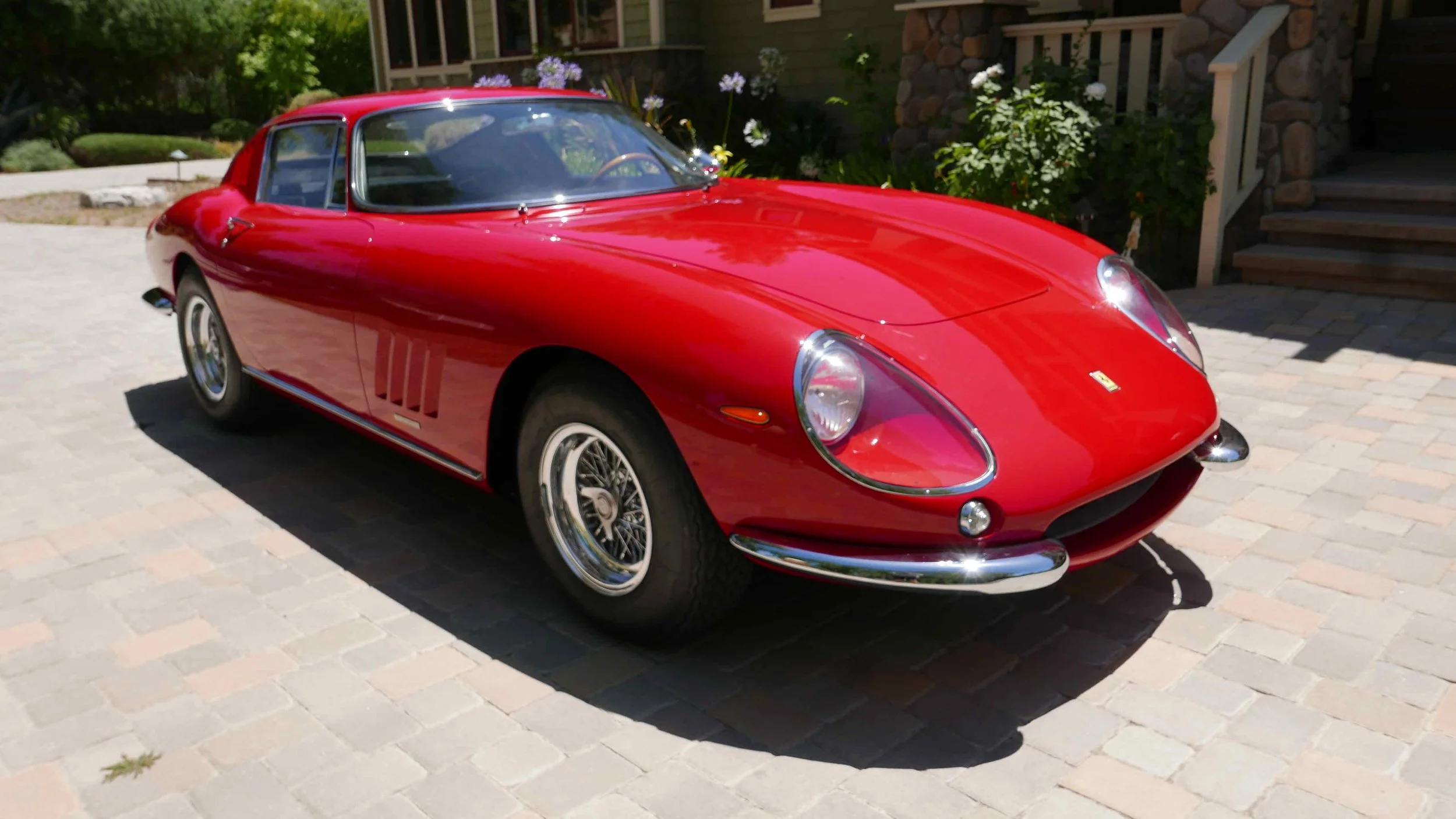 A red vintage sports car parked on a brick driveway in front of a house with stairs, plants, and flowers.