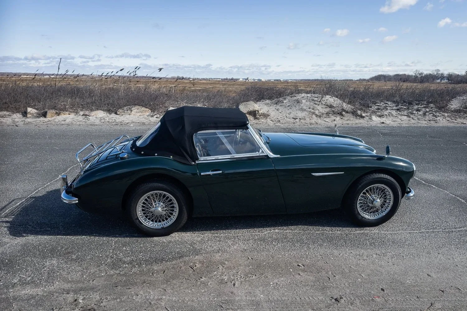 A vintage dark green convertible sports car with a black soft top parked on an empty roadside with a flat, open landscape and partly cloudy sky in the background.