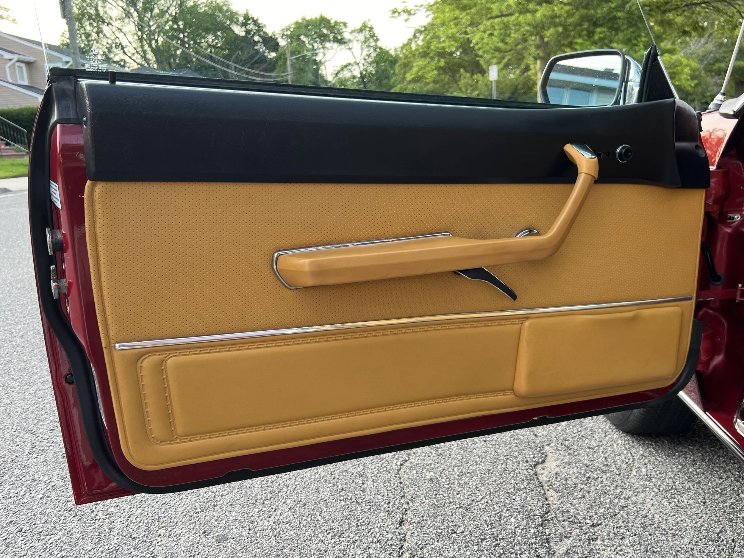 Close-up of a vintage 1977 BMW 630 csi car door interior with tan upholstery, black upper section, and chrome accents, parked on a street with trees in the background.
