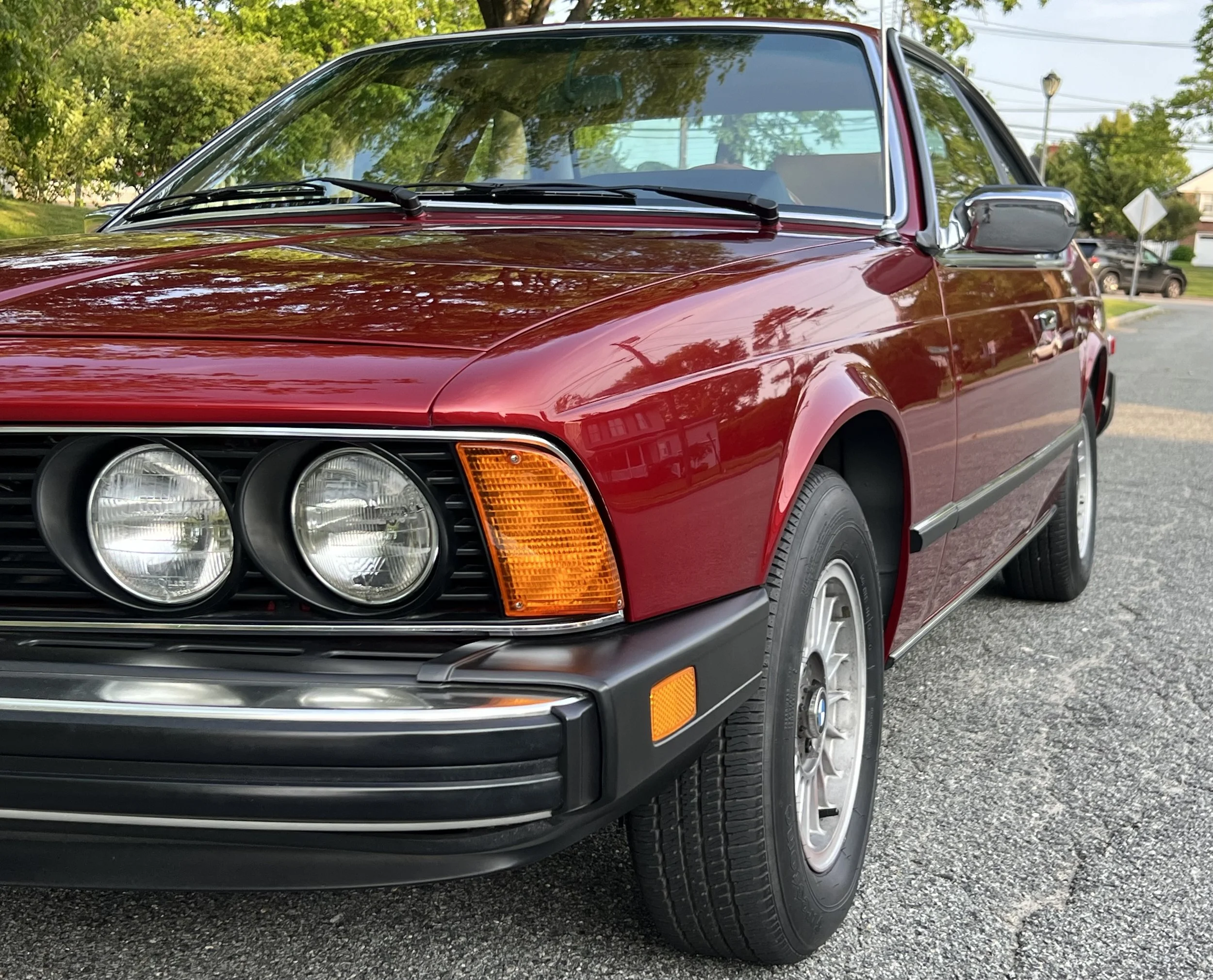 A red vintage BMW 630 csi car parked on a street with trees and houses in the background.