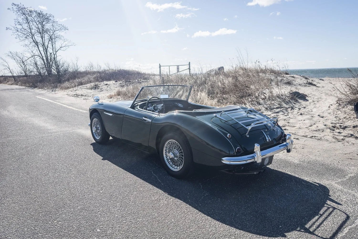 A vintage black convertible sports car parked on the side of a coastal road with sand dunes, dry grass, a few trees, and the ocean in the background.
