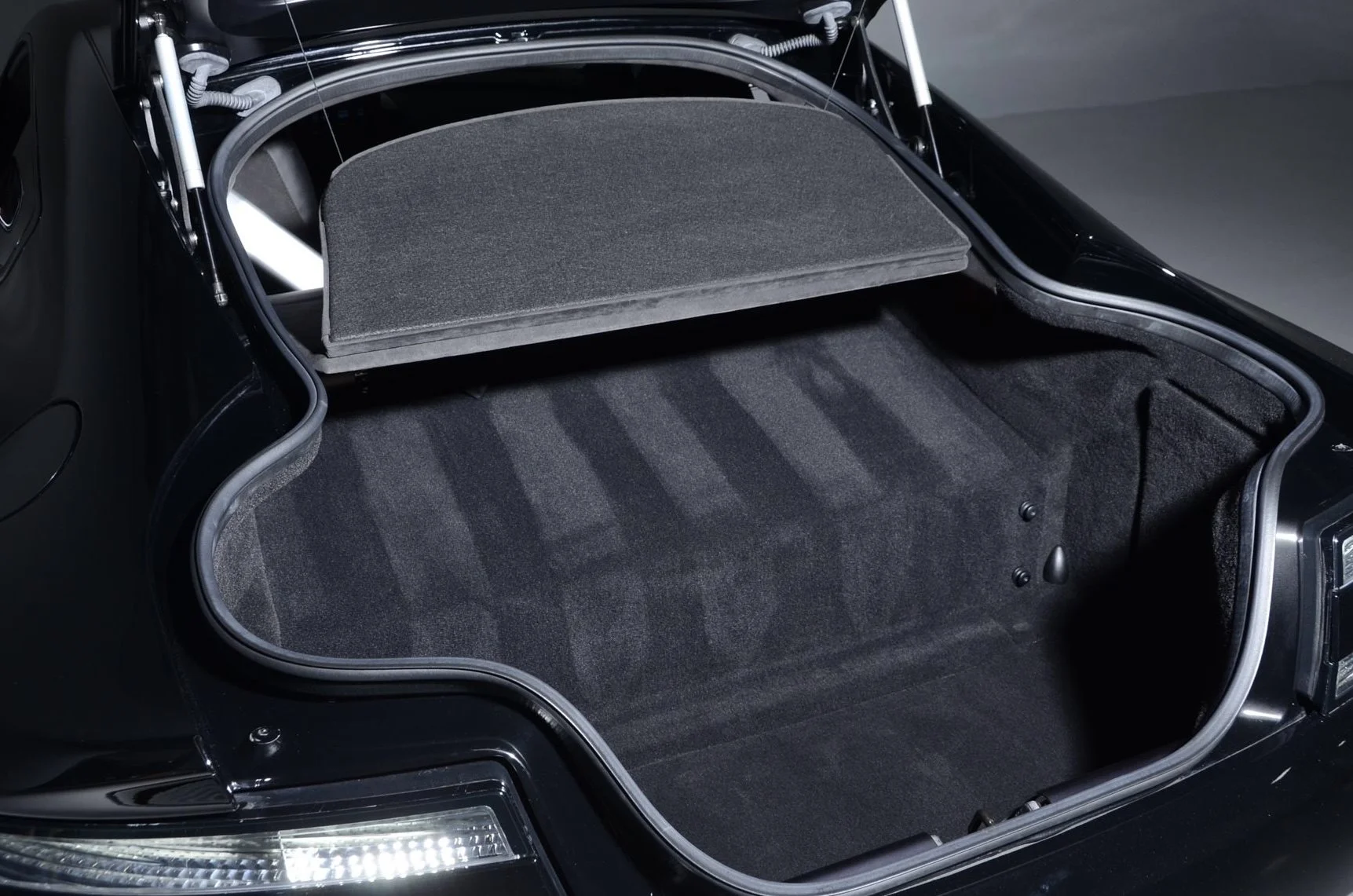 The trunk of a black sports car, with the trunk lid open, showing a spacious interior with dark carpeted flooring and a panel covering the spare tire area.