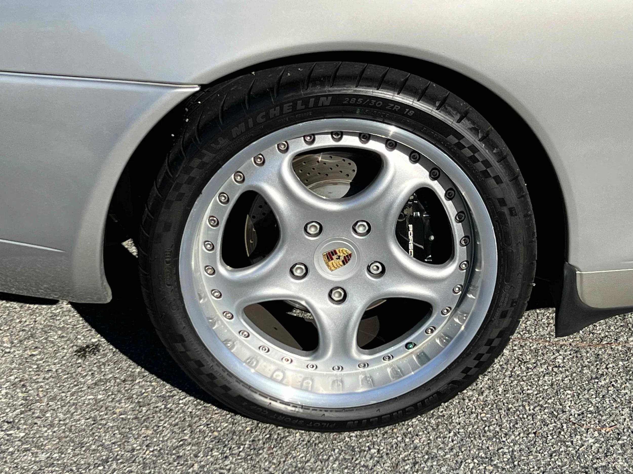 Close-up of a silver Porsche car wheel with a racing-style tire and a brake caliper visible through the rim.