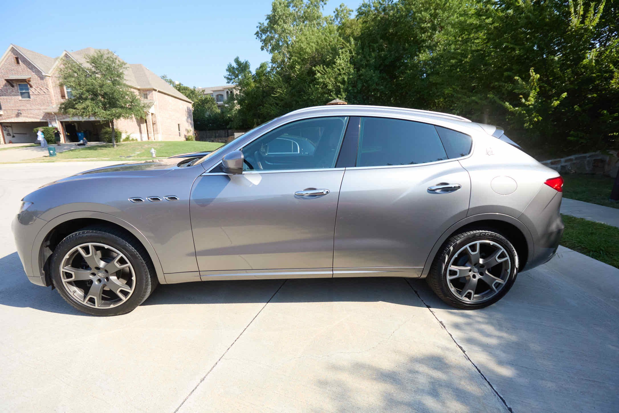 Silver luxury SUV parked on a driveway in front of a suburban house with trees and clear sky in the background.