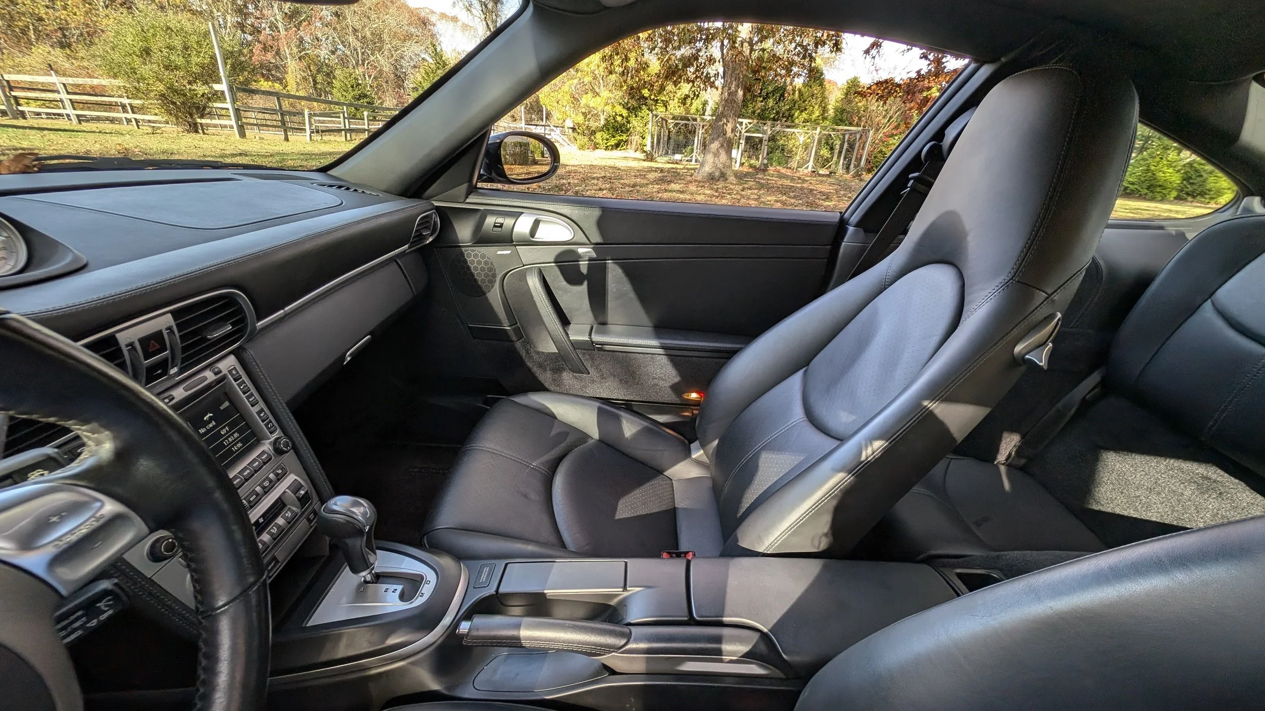 Interior of a sports car with black seats, gear shift, dashboard, and a view of outdoor trees and fences through the window.