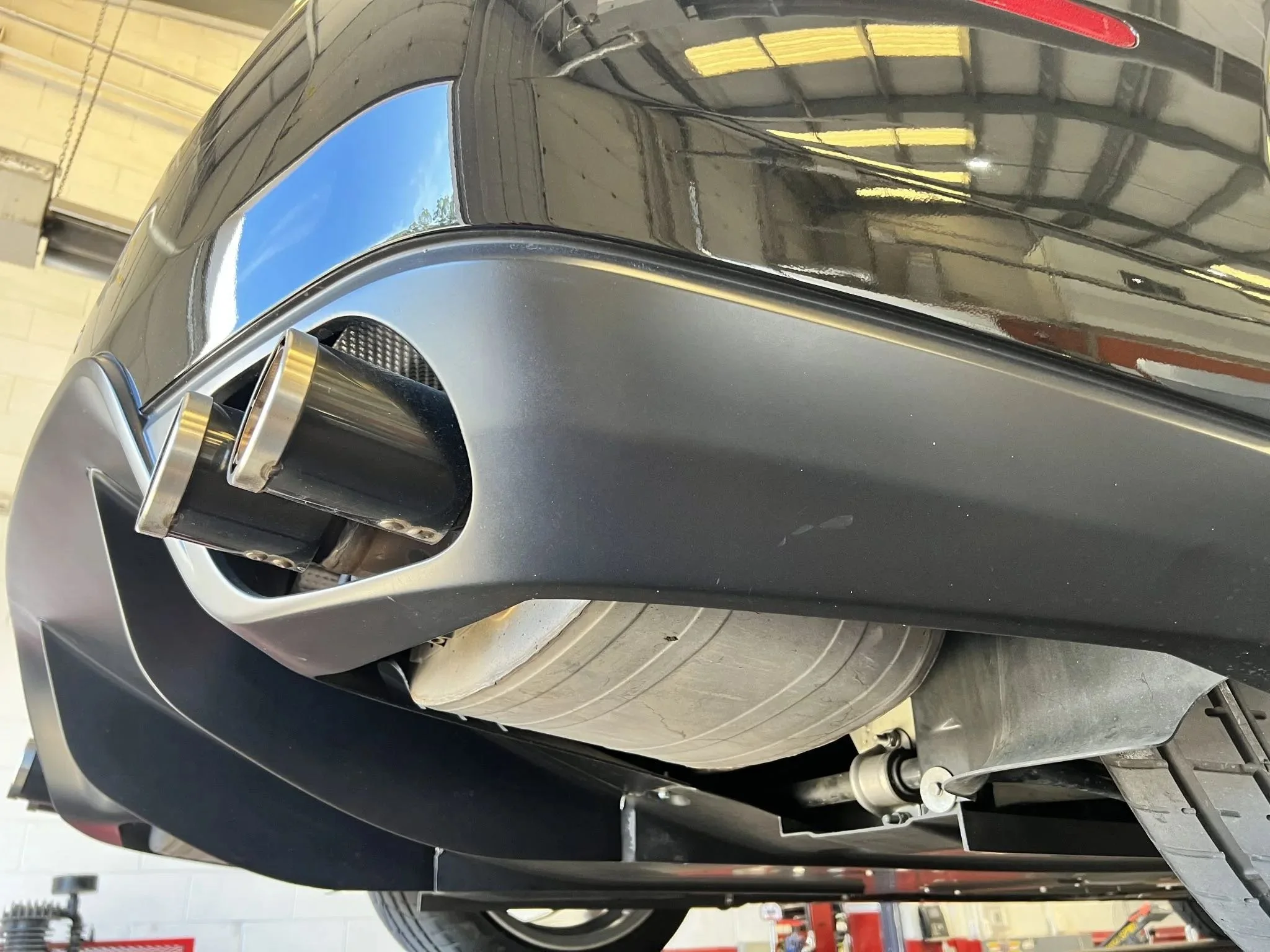 Close-up view of a car's exhaust pipe system attached to the rear of a black vehicle, with a large metallic muffler and dual exhaust tips.