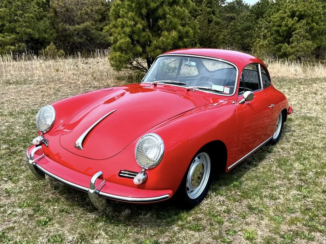 A vintage red sports car parked on grassy field with trees in background.