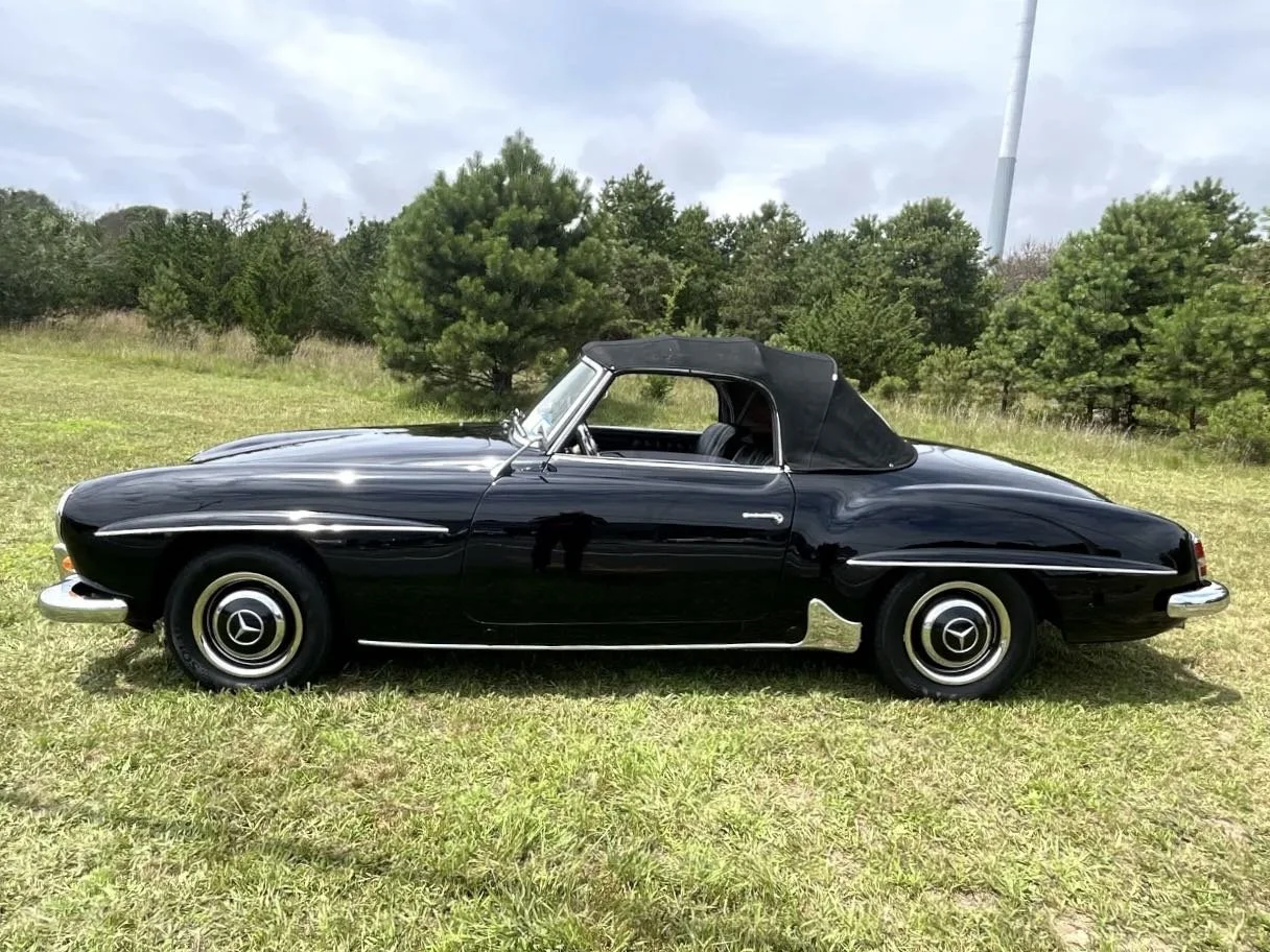 Black vintage Mercedes-Benz convertible car parked on grass with trees and cloudy sky in the background.