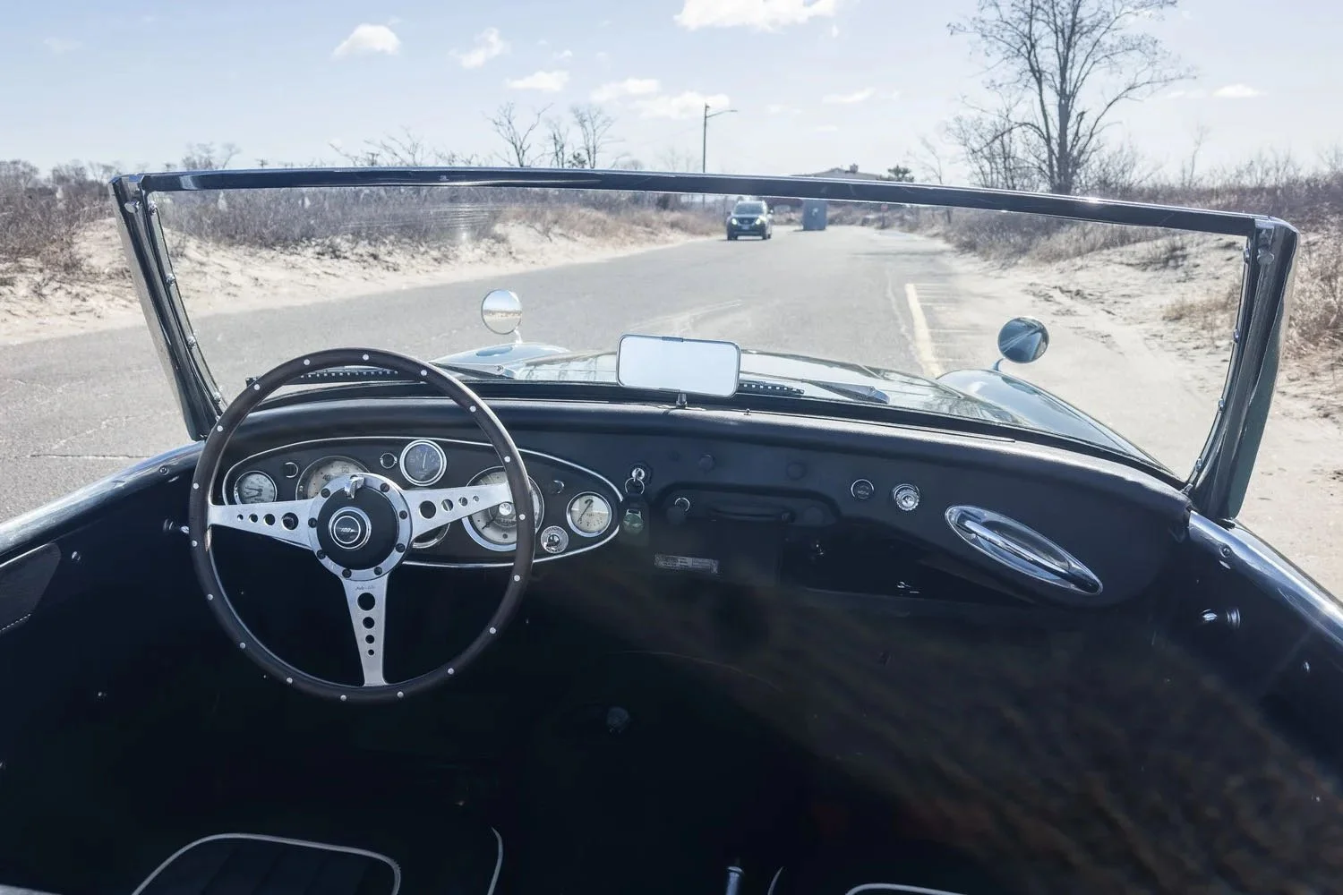 View from inside the vintage open-top car showing the dashboard, steering wheel, and rural road with scattered cars, sand dunes, and leafless trees in the background.