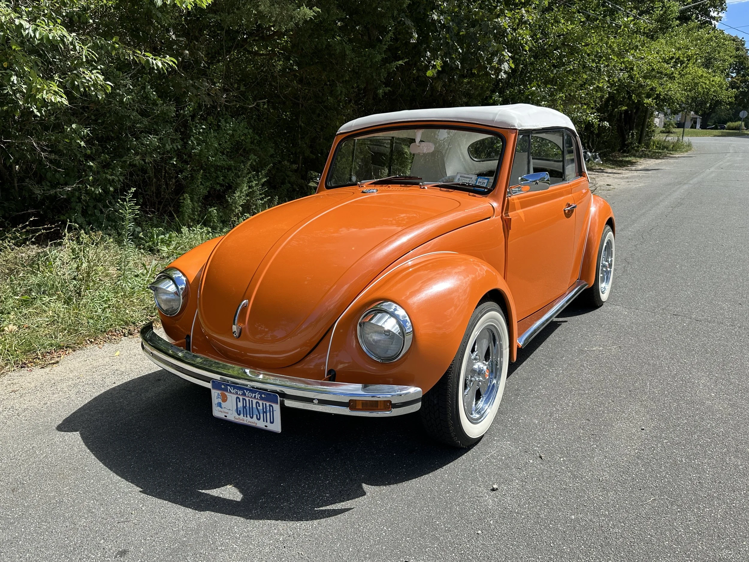 An orange vintage Volkswagen Beetle convertible parked on the side of a road with green bushes and trees in the background, and a New York license plate that reads "GRUSH".