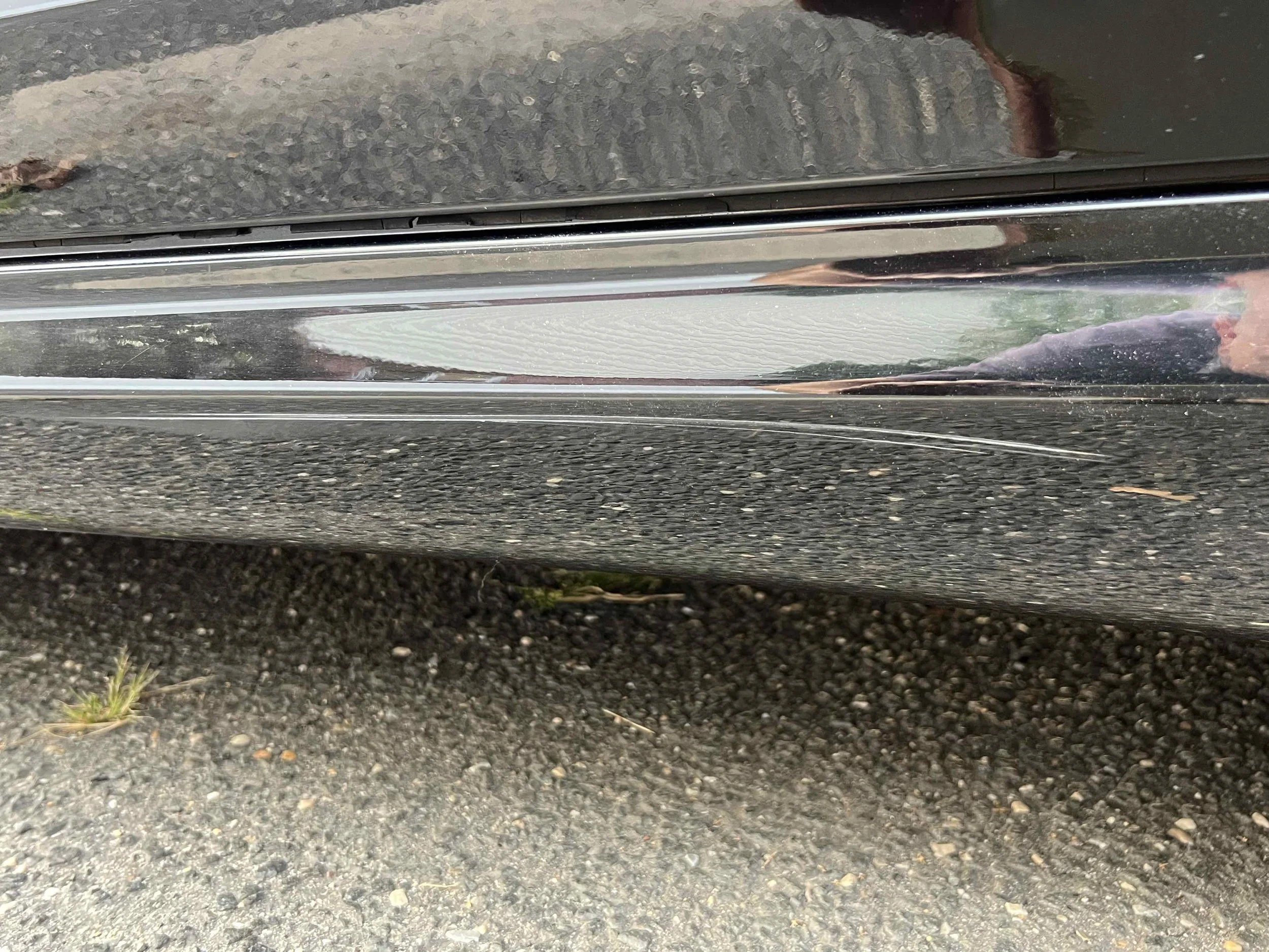 Close-up of a black car door with a visible long scratch on the lower part, parked on a gravel surface.