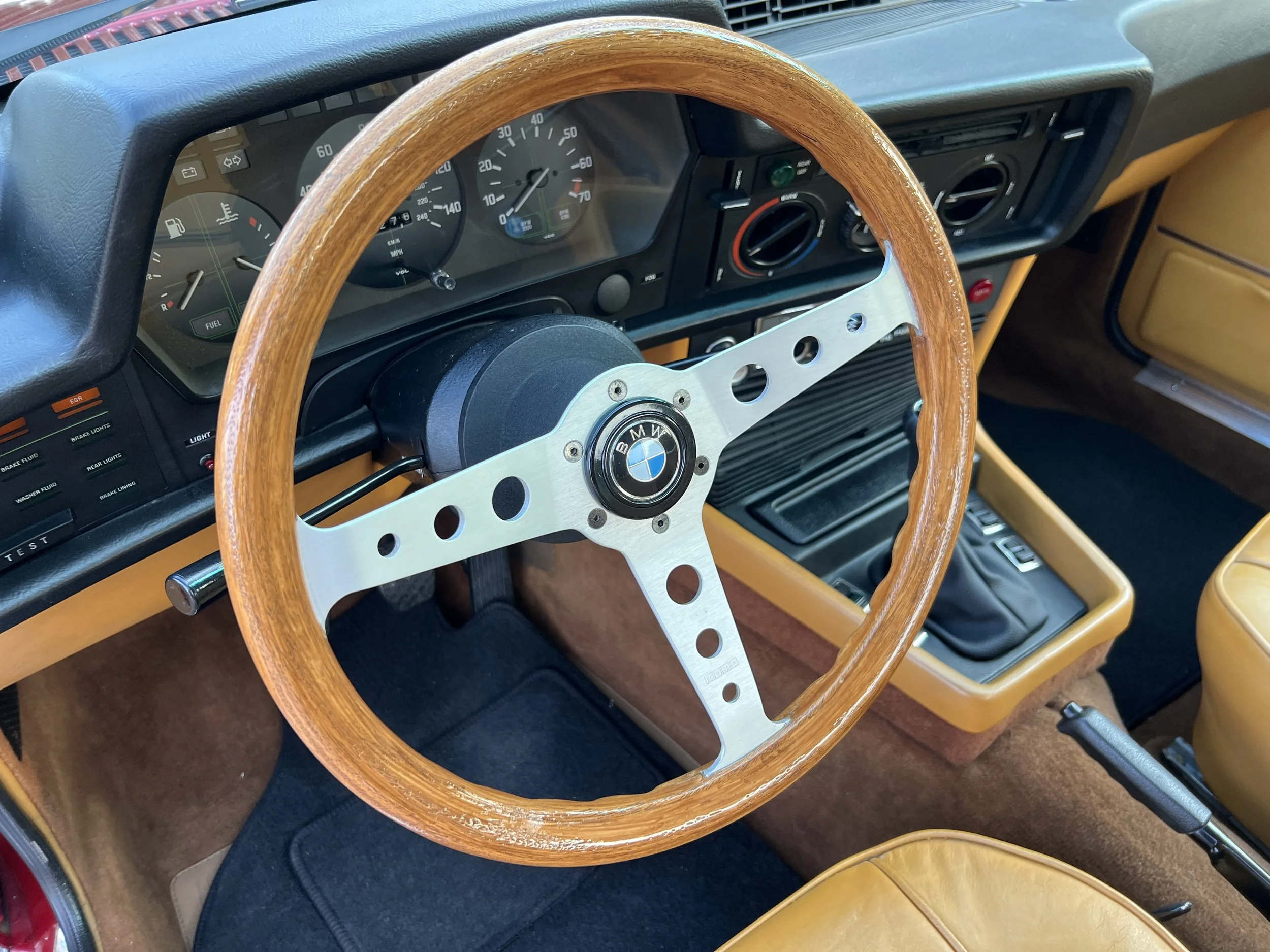 Close-up of the interior of a vintage BMW BMW 630 csi car, showing a wooden steering wheel, dashboard with gauges, and tan leather seats.