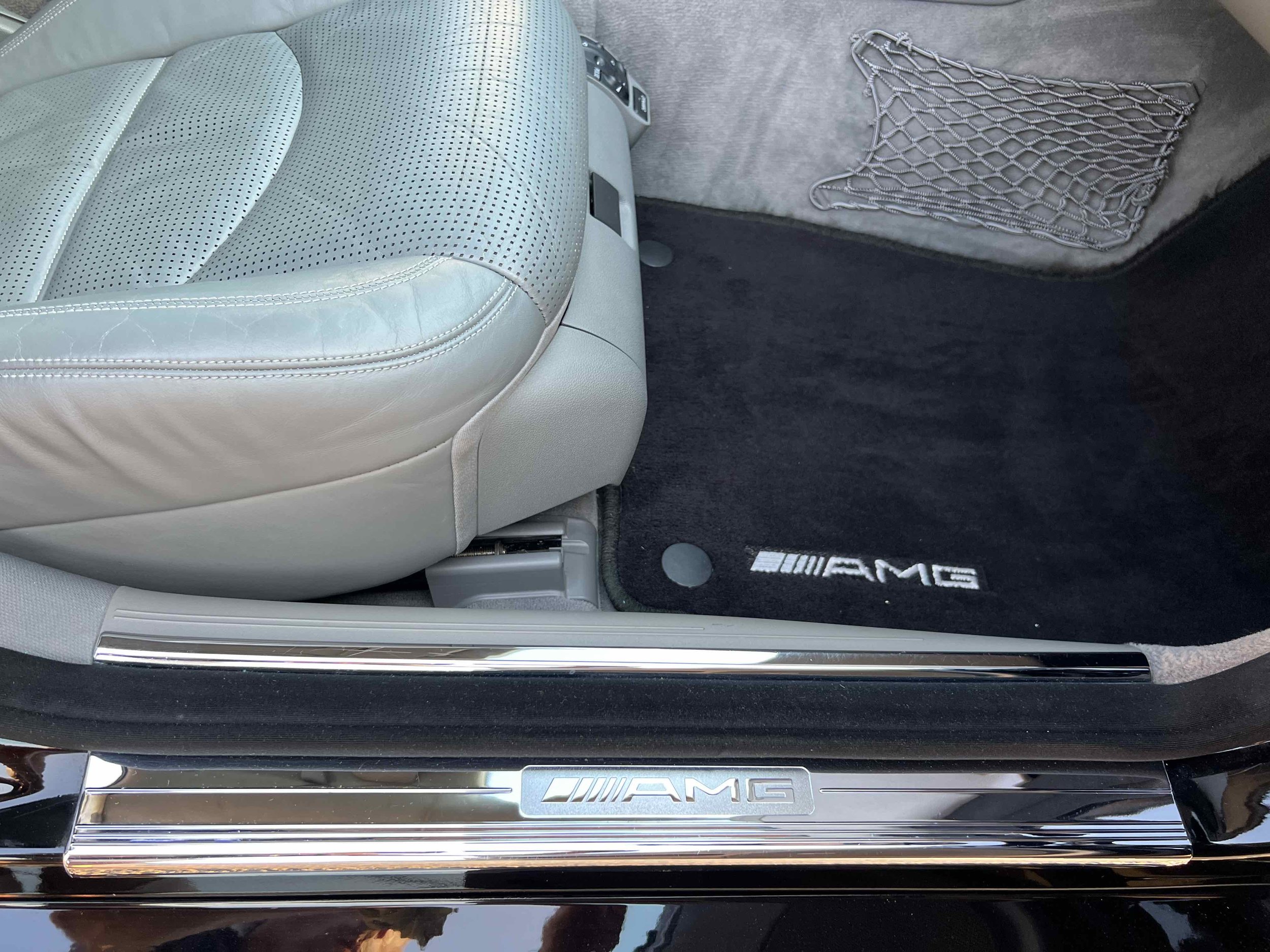 Close-up of the interior of a Mercedes-Benz AMG vehicle, showing a light gray leather seat, a black carpeted floor mat with the AMG logo, and a silver AMG-branded door sill plate.