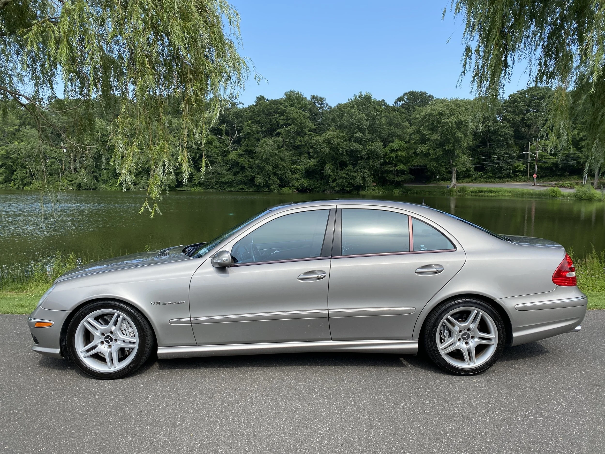 Side view of a silver Mercedes-Benz E-Class sedan parked on the side of a road near a lake with trees and blue sky in the background.