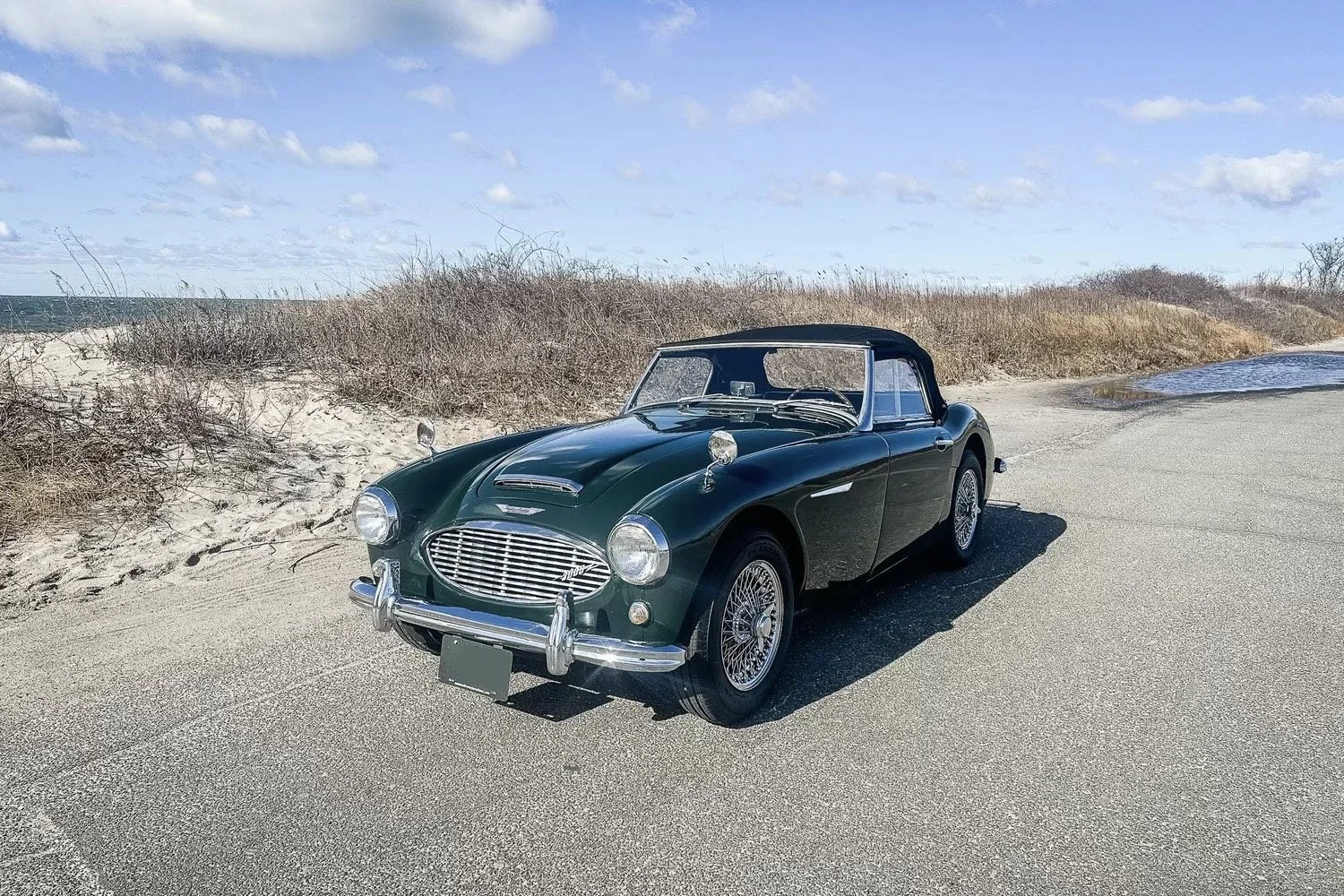 A vintage dark green convertible sports car parked on a sandy beachside road with dunes and sparse vegetation in the background, under a partly cloudy sky.