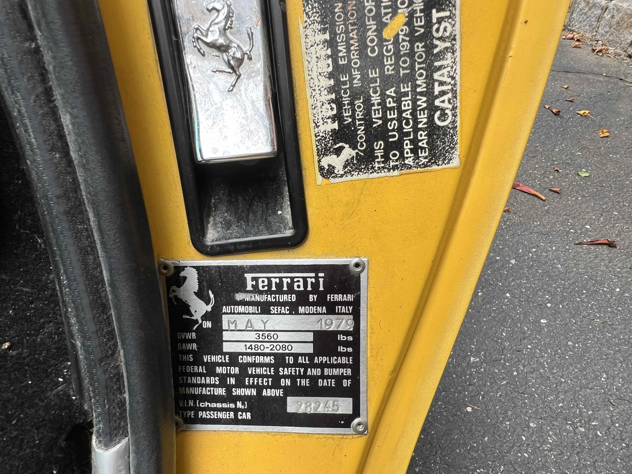 Close-up of a yellow vehicle identification plate from a Ferrari, showing it was manufactured in May 1970 in Modena, Italy, with a VIN of 28245. The plate includes technical details such as GVWR and GAWR.