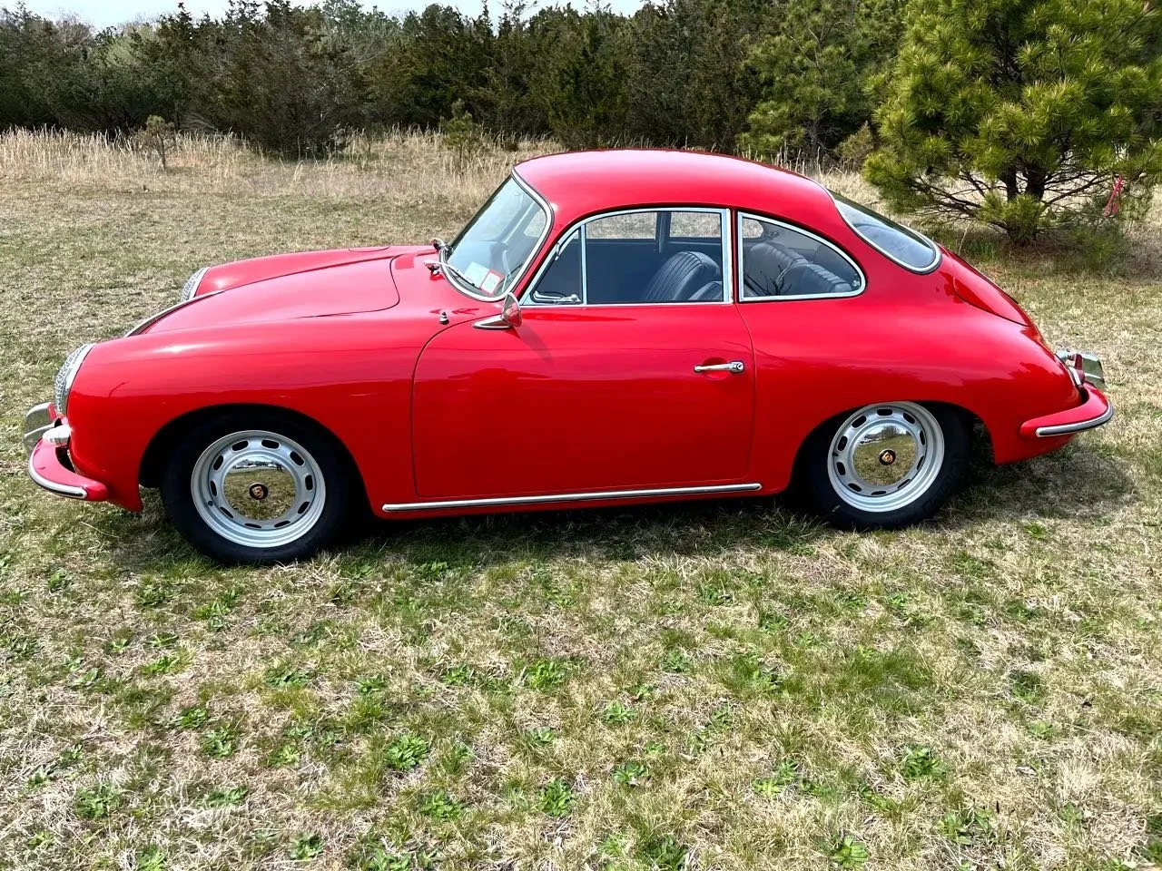 Red vintage Porsche 356 parked on grassy field with trees in background.
