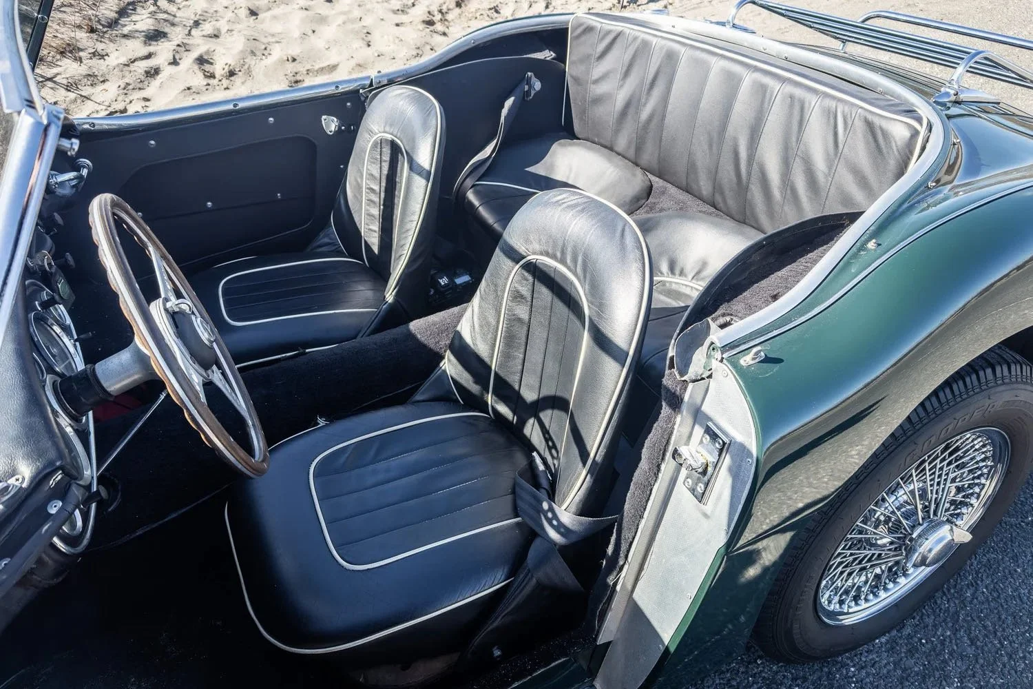 Interior of a classic green convertible car with black leather seats and wire spoke wheels, parked on a sandy surface.