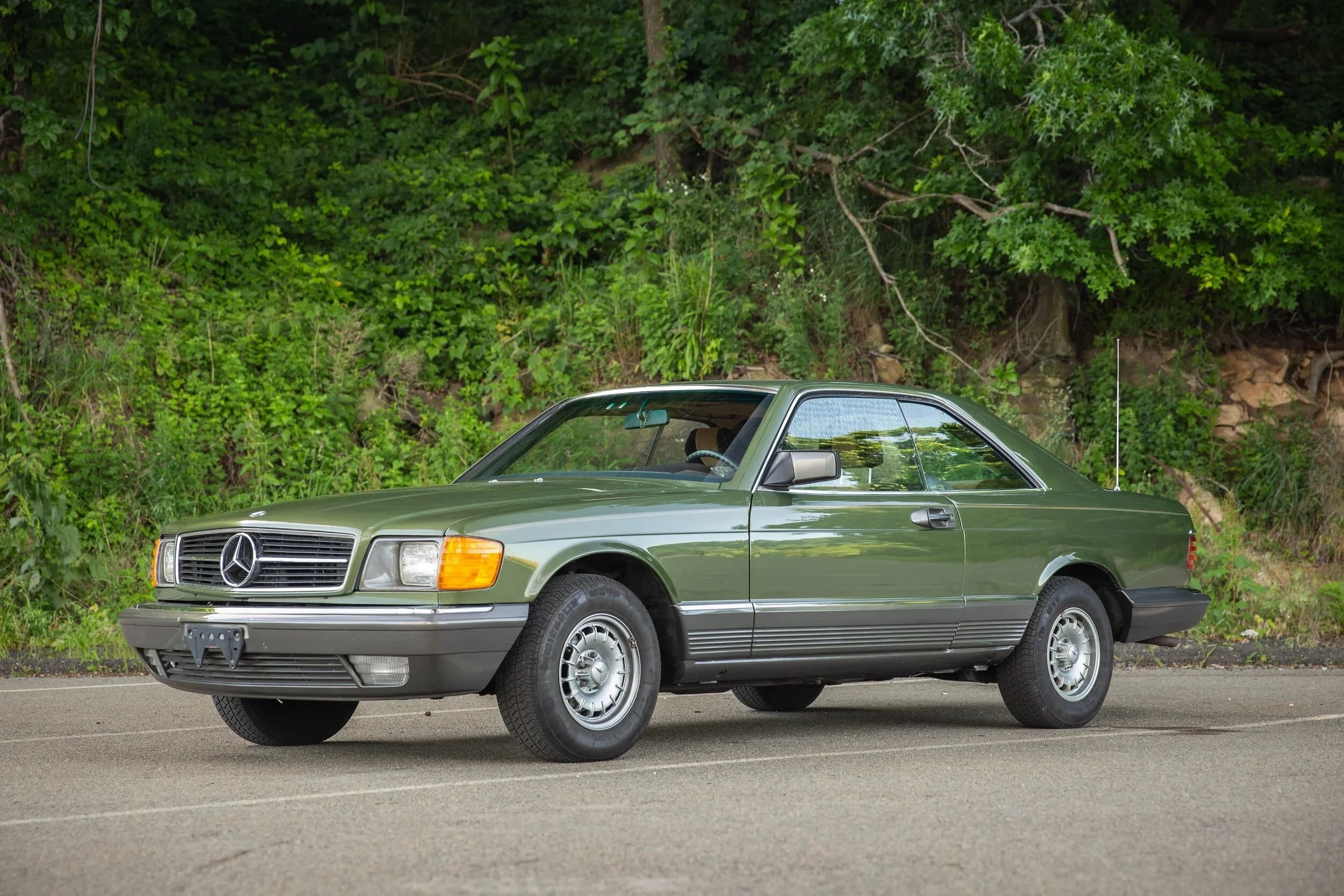 A green vintage Mercedes-Benz coupe parked on the side of a paved road with green foliage in the background.