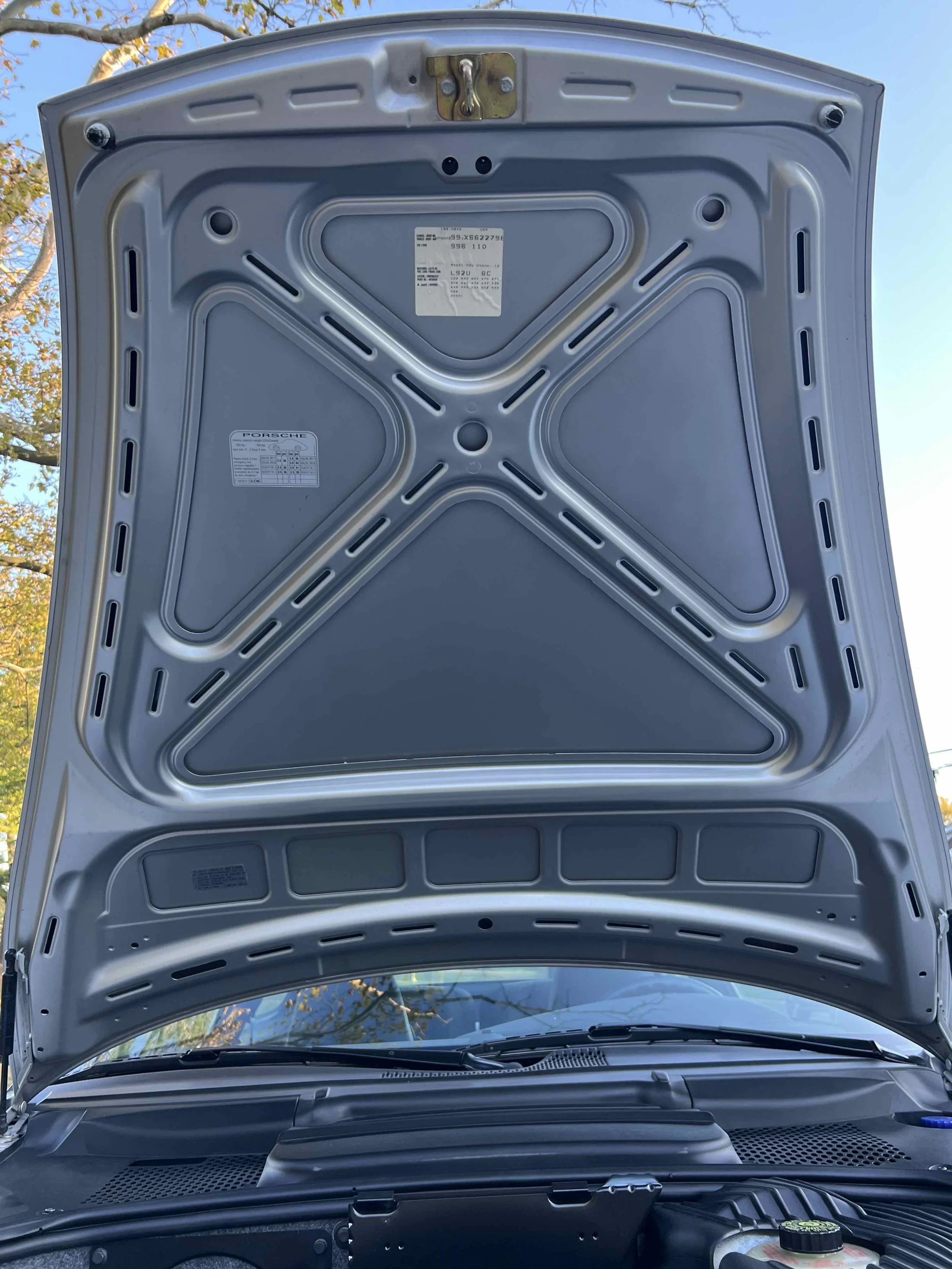 The image shows the underside of a car's open hood, revealing the metal structure and some labels, with trees and a clear sky in the background.
