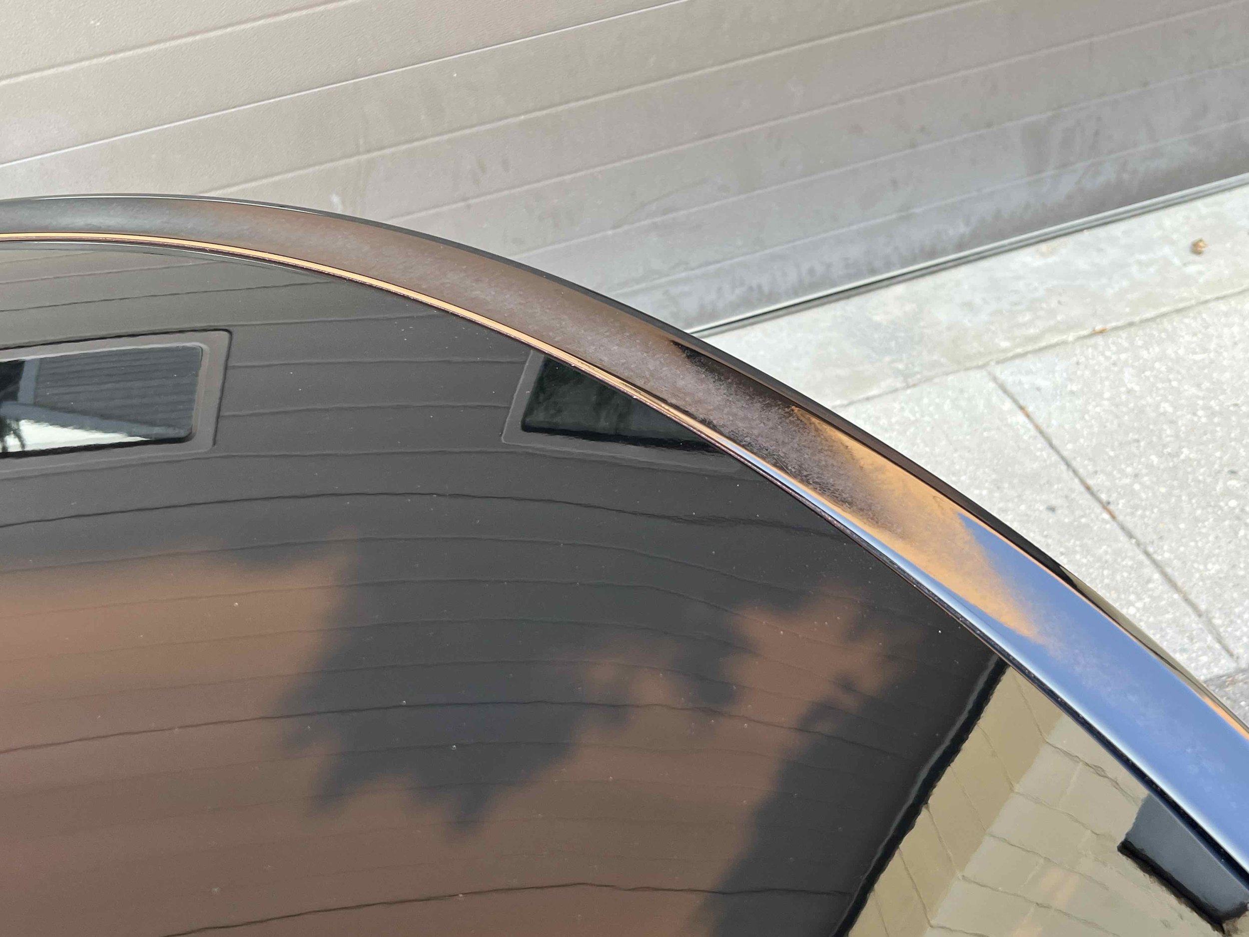 Close-up of a glossy black car roof with a metallic trim, reflecting a cloudy sky and nearby structures.