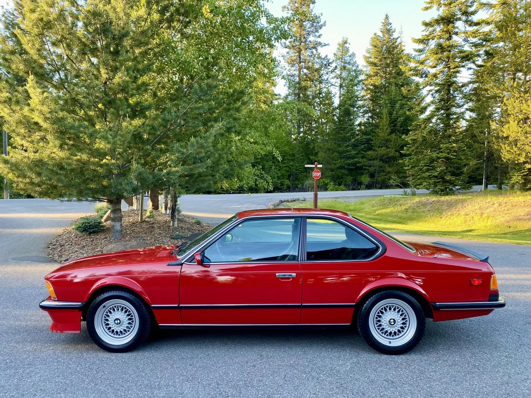 A red vintage two-door coupe car parked on a paved road with trees and a stop sign in the background