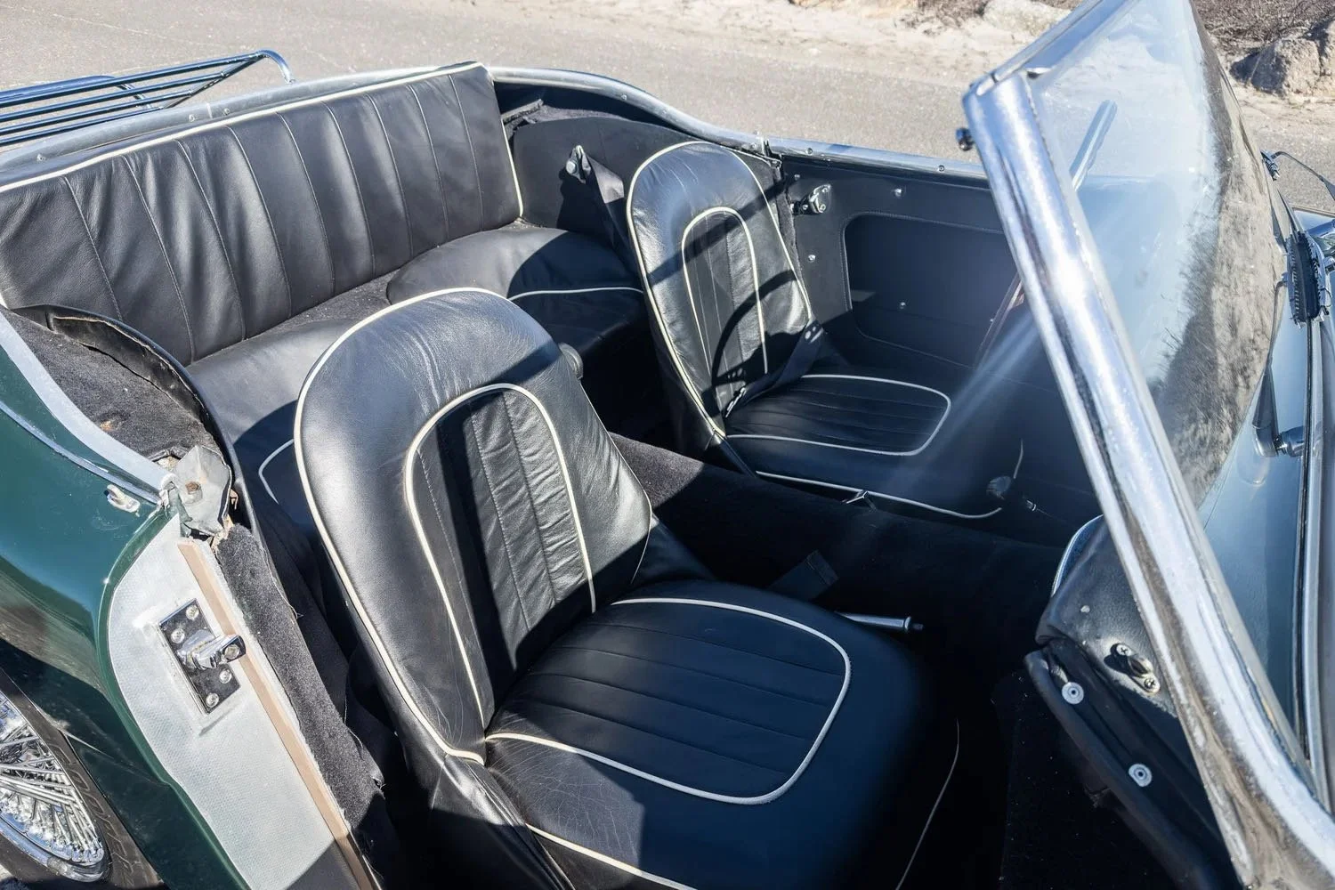 Interior of a vintage convertible car with black leather seats and a clear windshield, parked on a sandy or gravel surface.