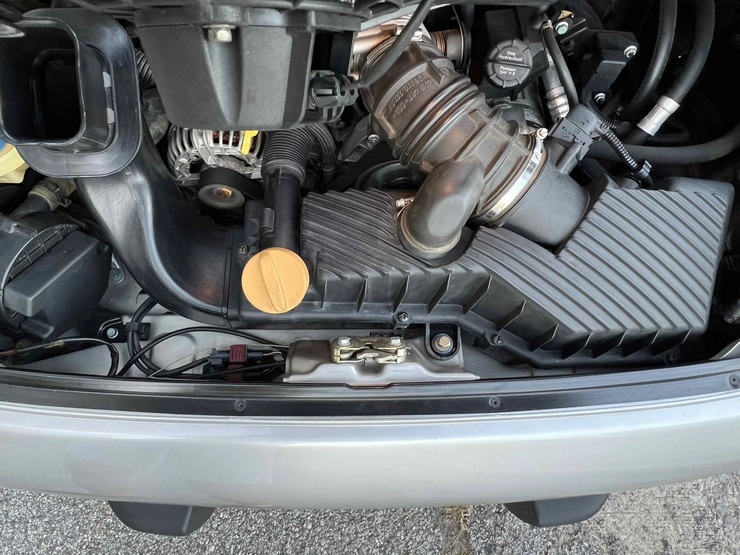 A car engine compartment showing various components, including a large black intake manifold, an orange oil cap, a black radiator hose, an alternator, and other engine parts.