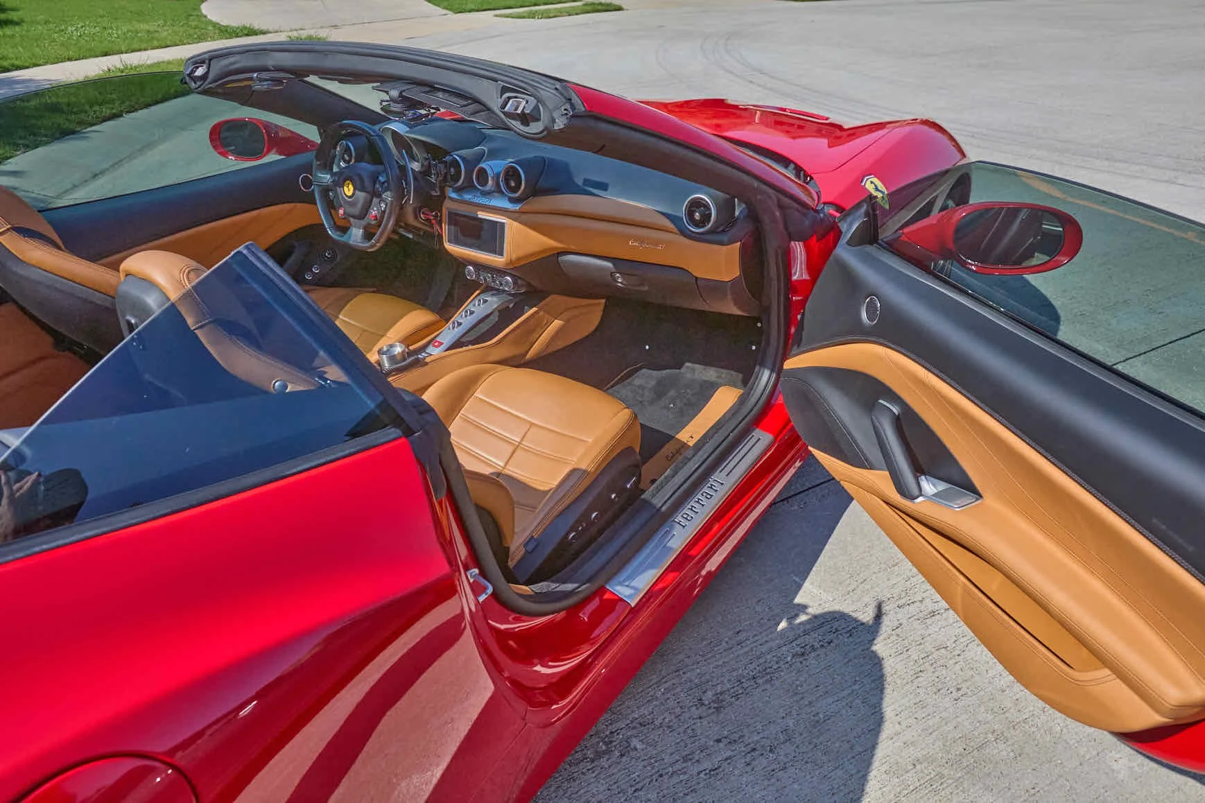 Red convertible sports car with tan leather interior and dashboard, parked on a driveway with the driver's side door open.