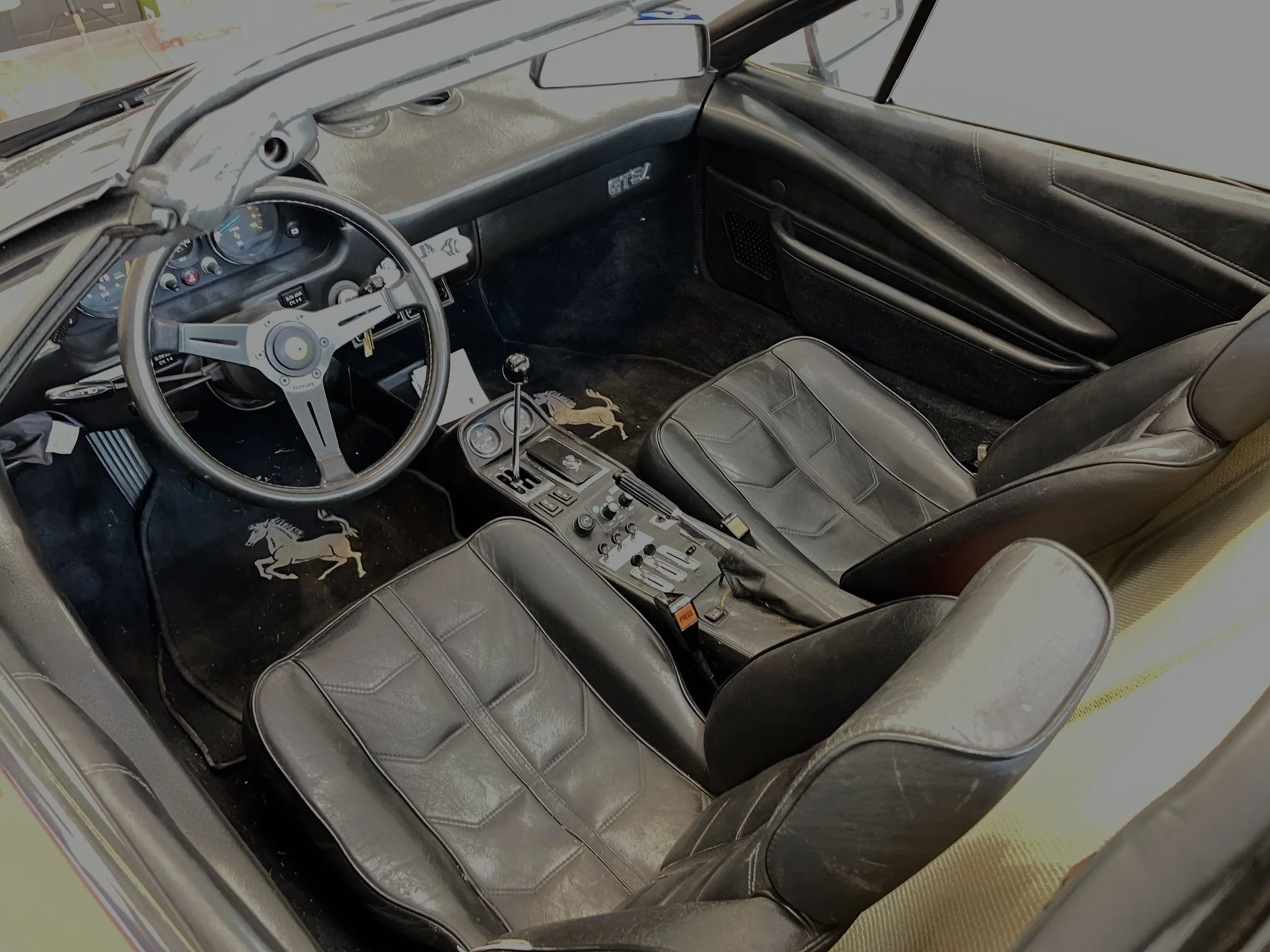 Interior of a classic race car with black leather bucket seats, a dashboard with gauges, and a central gear shifter, showing signs of wear and a Ferrari logo on the floor mats.