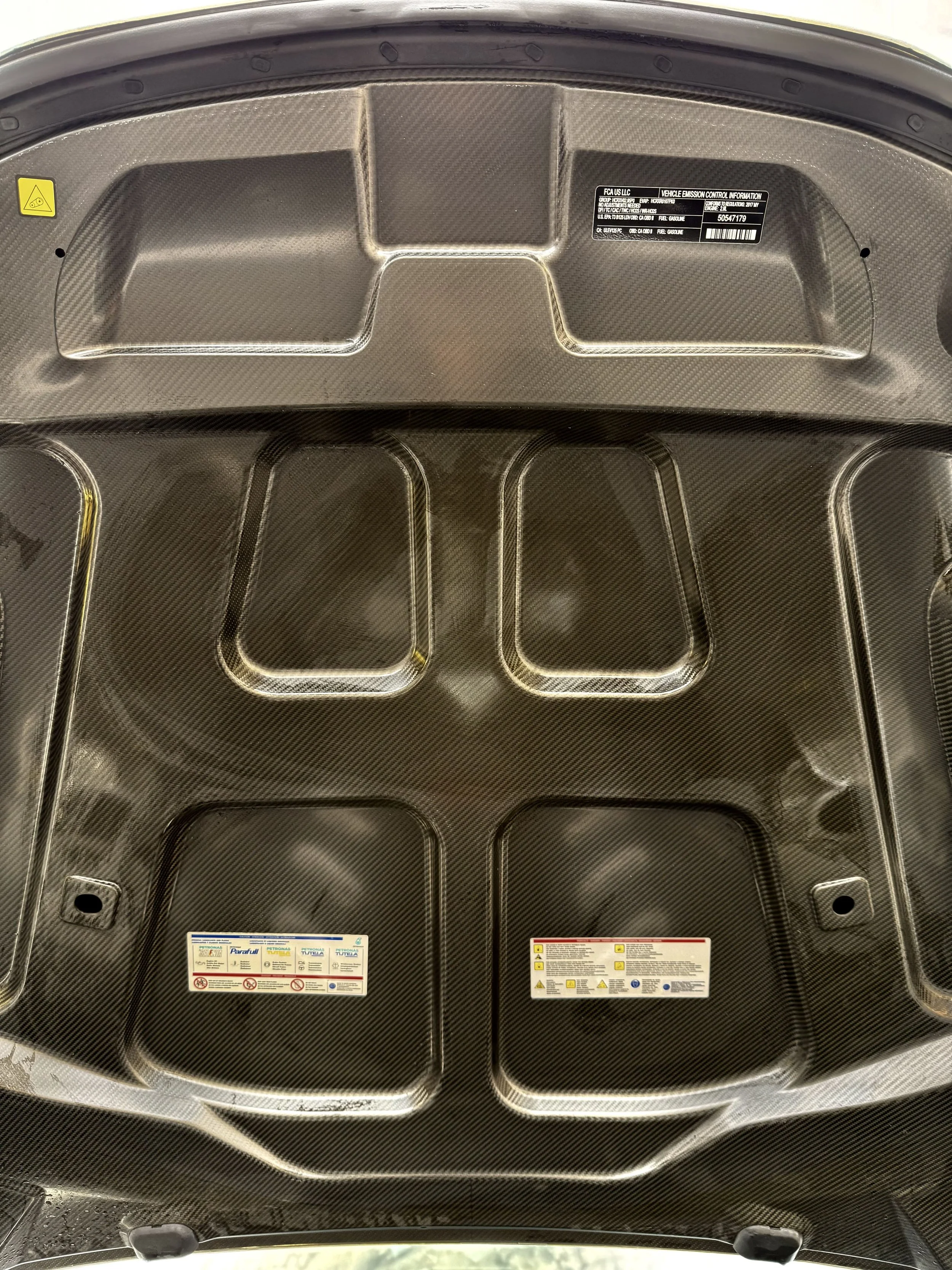 The underside of a car hood made of carbon fiber with warning labels and vehicle emission control information.