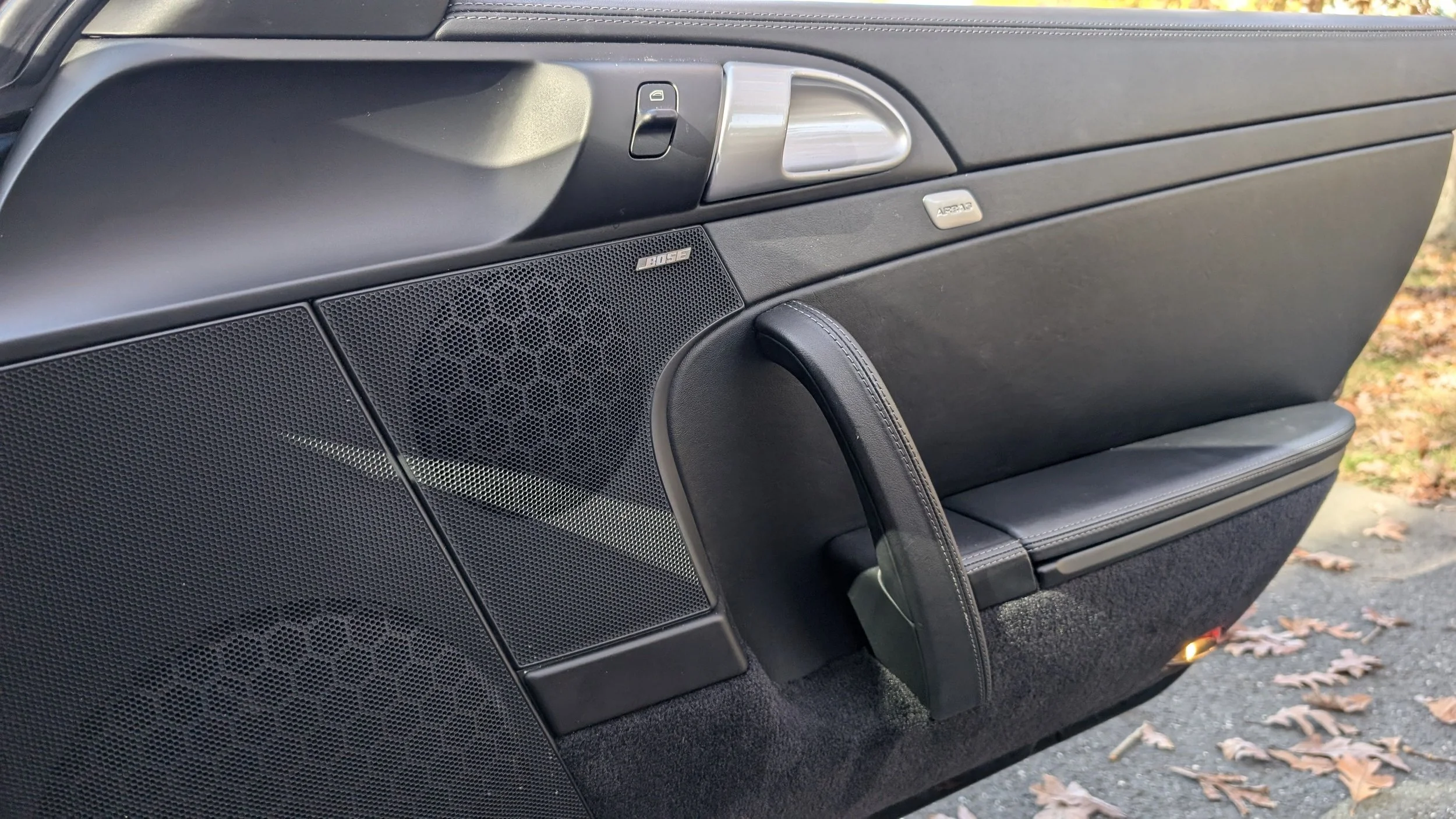 Close-up of the interior of a car's passenger side door panel, showing a speaker with a Bosch logo, a window control button, a door handle, and a small orange light near the floor. Outside the car, fallen autumn leaves are visible on the ground.