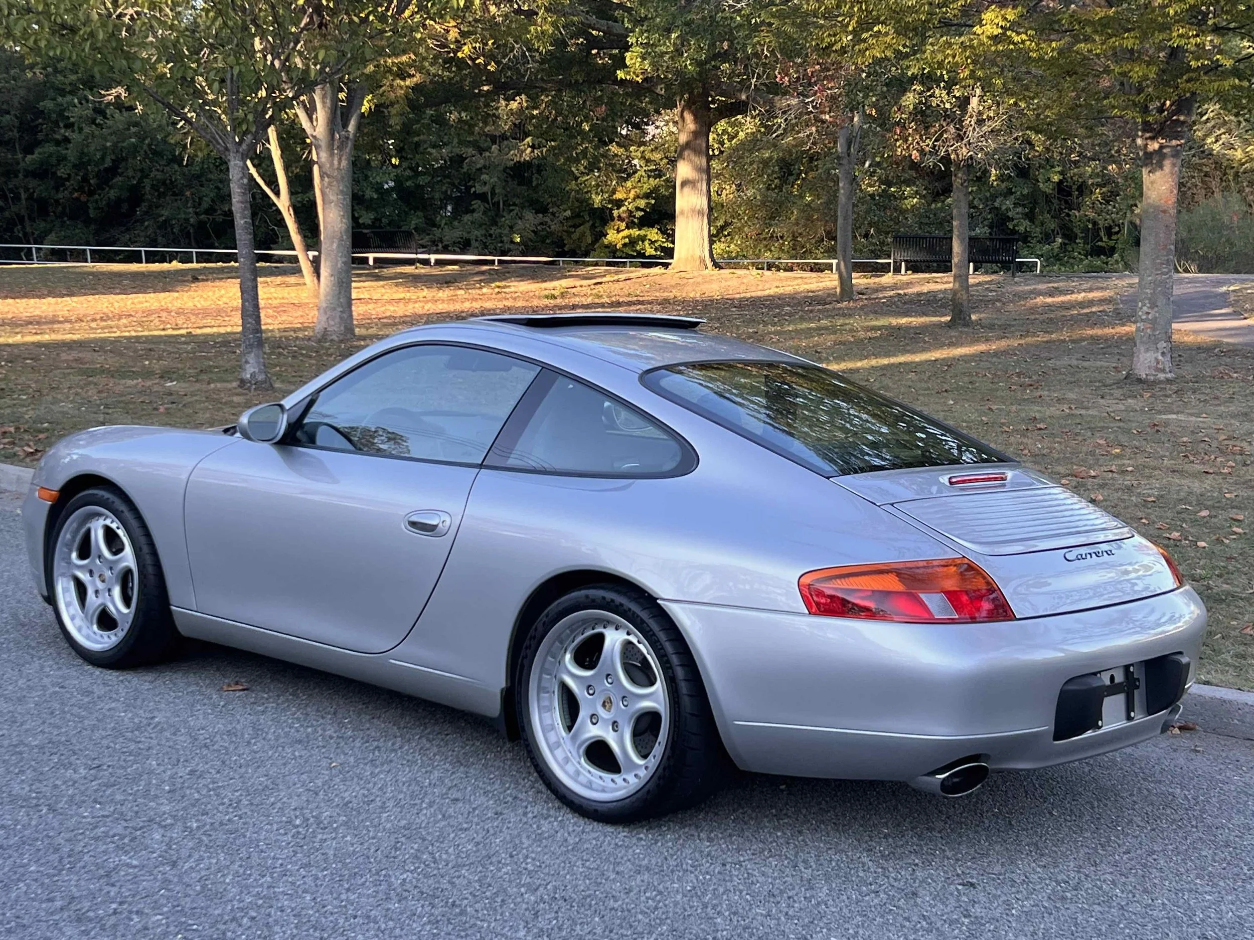 A silver Porsche Carrera parked on the side of a road, with trees in the background and shadows indicating late afternoon sunlight.