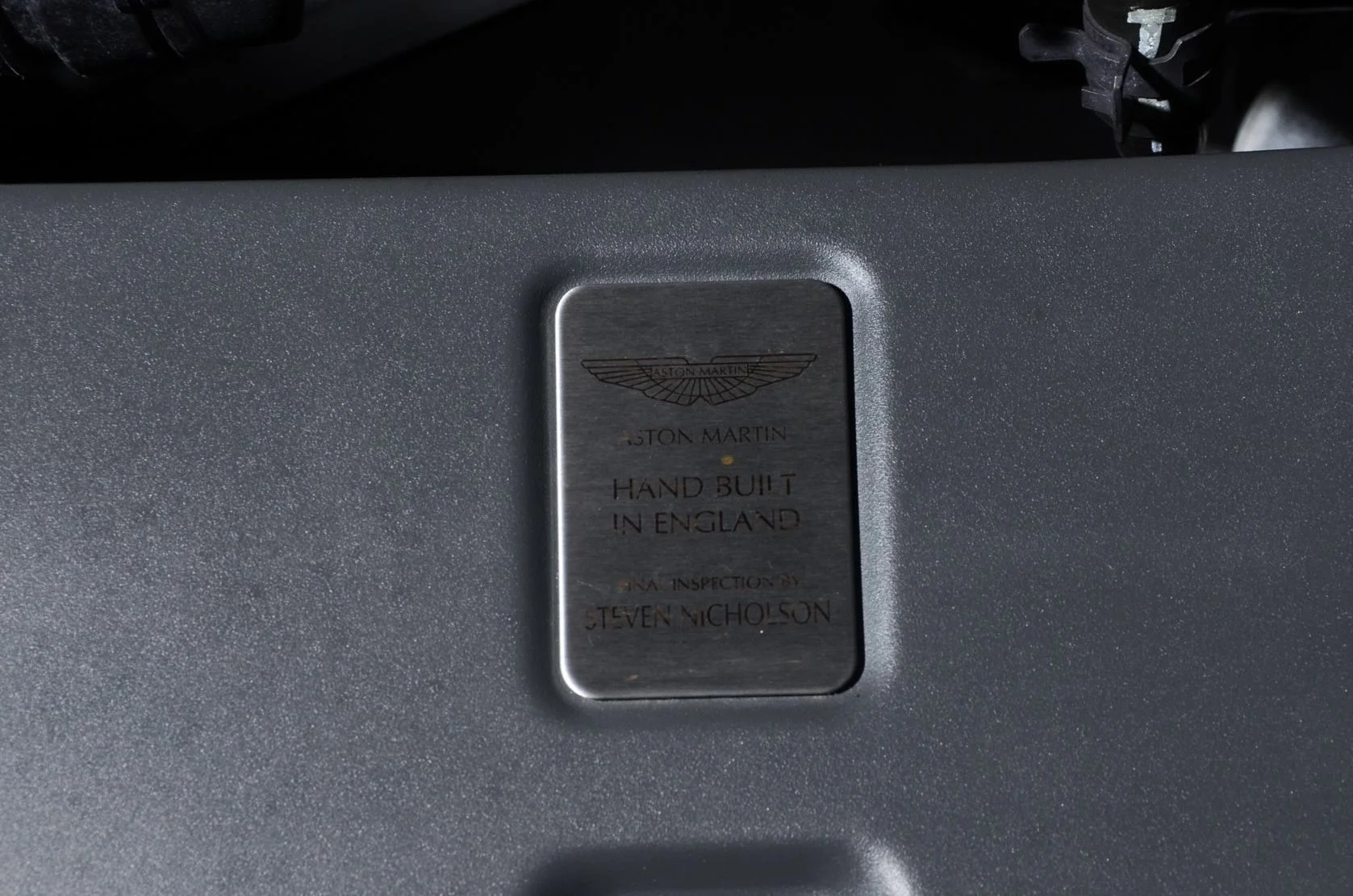 Metal plaque with Aston Martin logo and text, indicating the car was hand-built in England and inspected by Steven Nicholson.