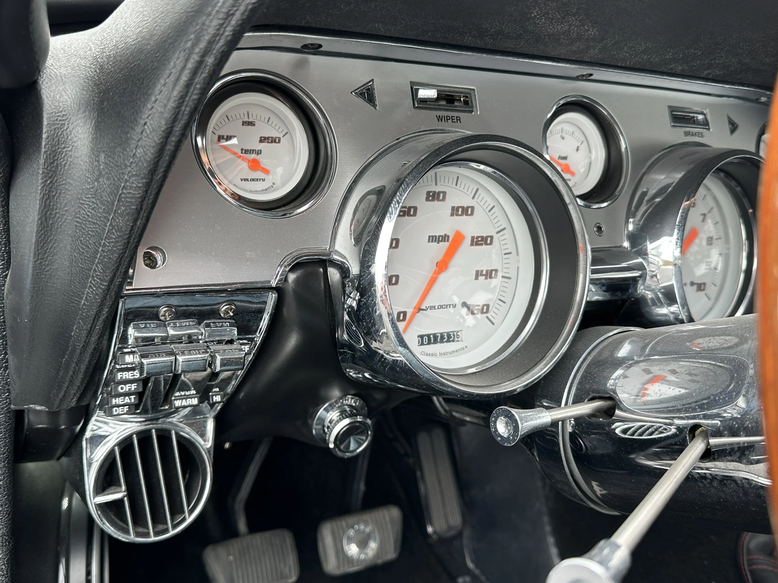 Close-up of a classic car dashboard with a white speedometer showing approximately 10 mph, and additional gauges displaying temperature, wiper, and brake information. There is a control panel with various switches on the left.