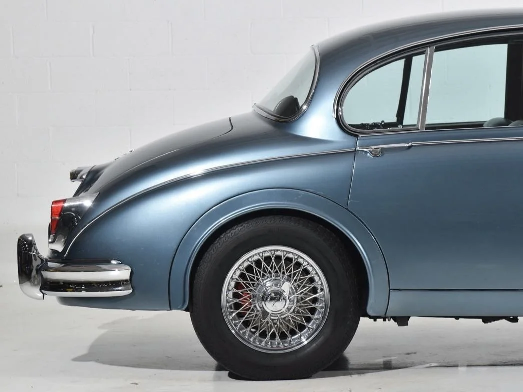 Close-up of the rear and side of a vintage blue car with wire-spoke wheel and chrome details, parked indoors with a plain white wall in the background.