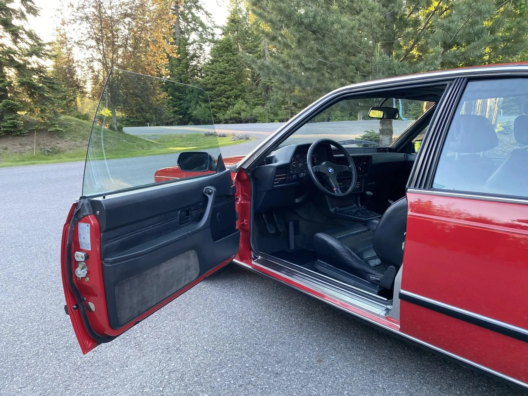 Open driver's side door of a red classic BMW car parked on a road, with the interior visible including black leather seats and dashboard, and a scenic background of trees and a curving road.