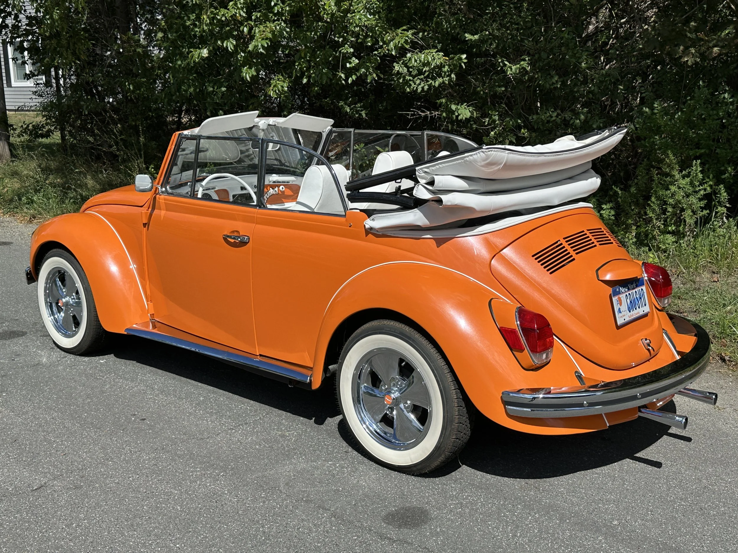 Orange vintage Volkswagen Beetle convertible with white interior and a folded white soft top, parked on a street with green trees in the background.