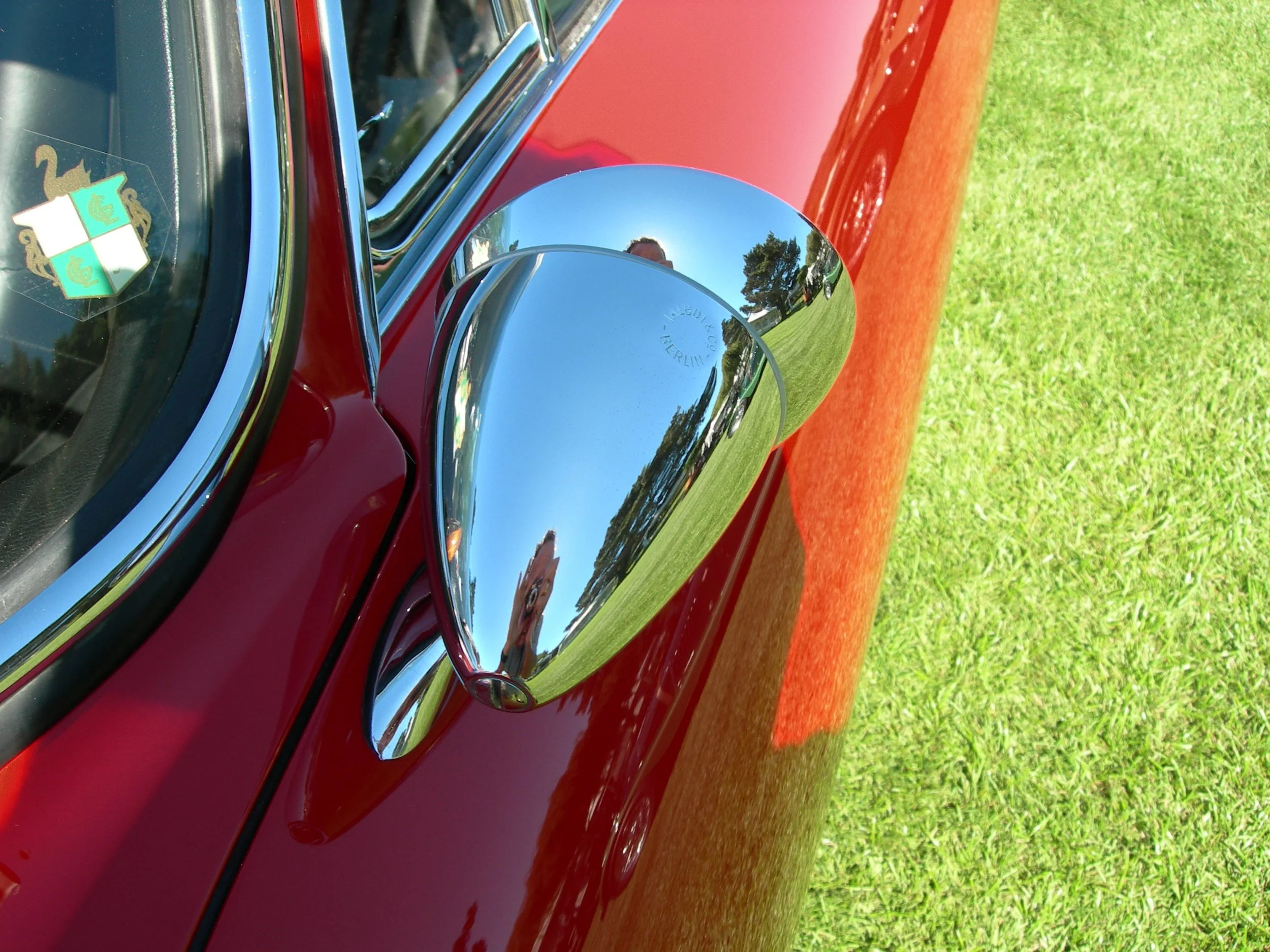 Close-up of red vintage car with shiny chrome side mirror and reflection of person, green grass, and trees in the mirror.