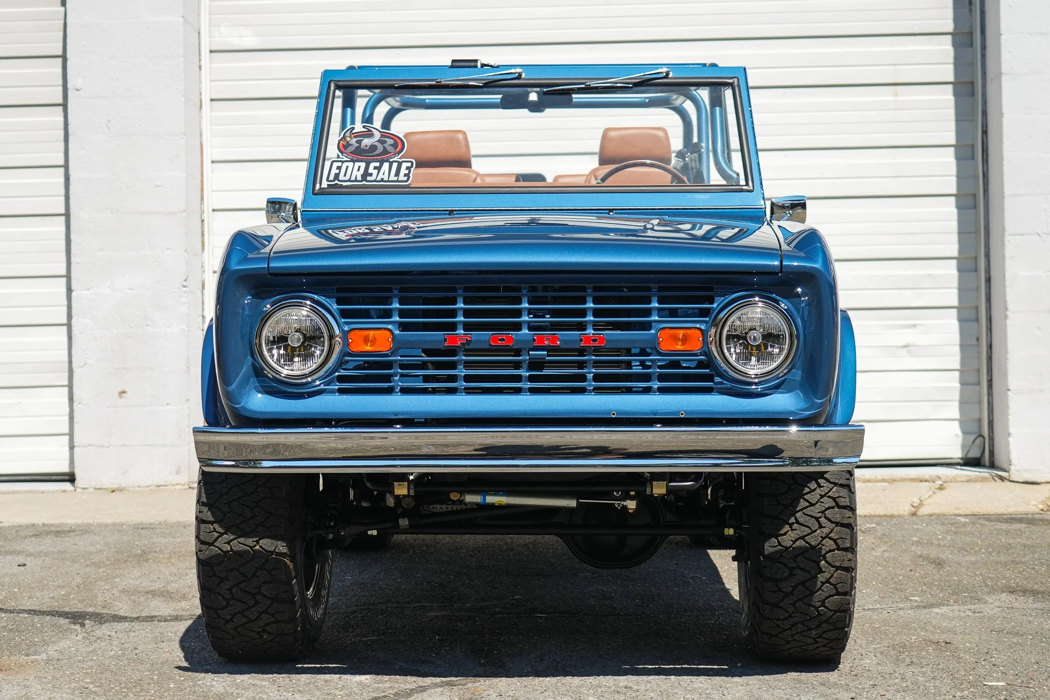 Front view of a vintage blue Ford truck with a 'For Sale' sign on the windshield, parked in front of white garage doors.