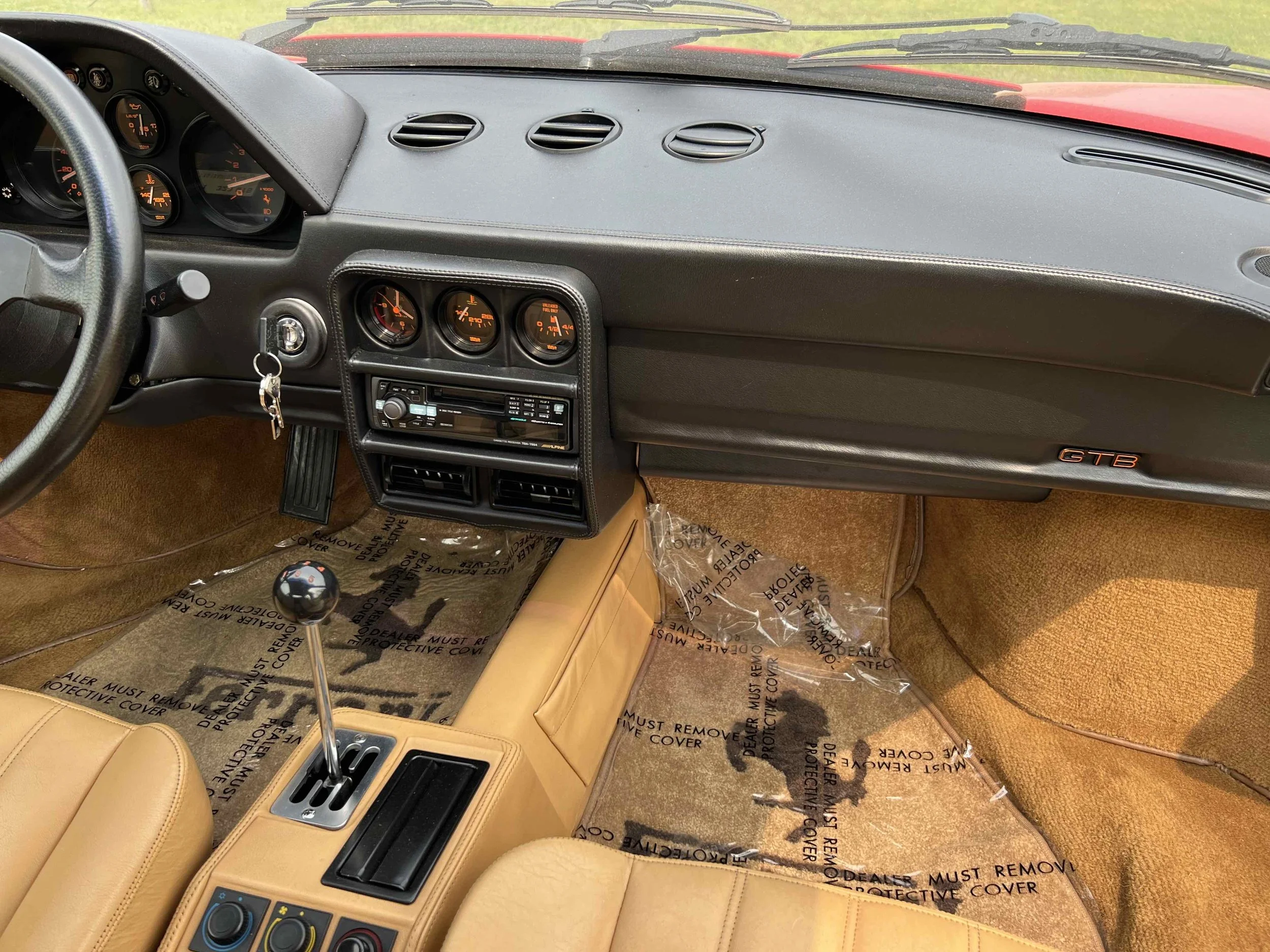 Interior of a classic Porsche 911 GTB, featuring a black dashboard with analog gauges, tan leather seats, a manual gear shift, and a protective plastic cover on the tan carpeted floor.