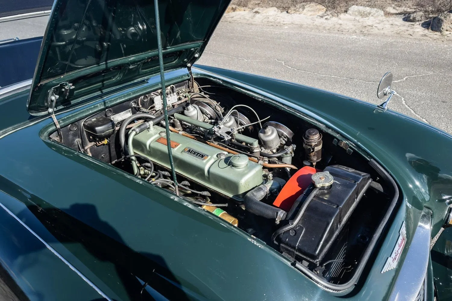 Vintage green car with its hood open, showing the engine compartment with a fuel intake, radiator, and various engine parts, parked on a paved surface.