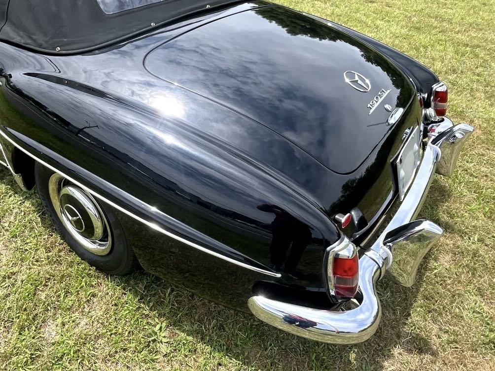 Rear view of a vintage black Mercedes-Benz 220SL convertible parked on grass, showcasing its chrome bumper, tail lights, and Mercedes-Benz emblem on the trunk.