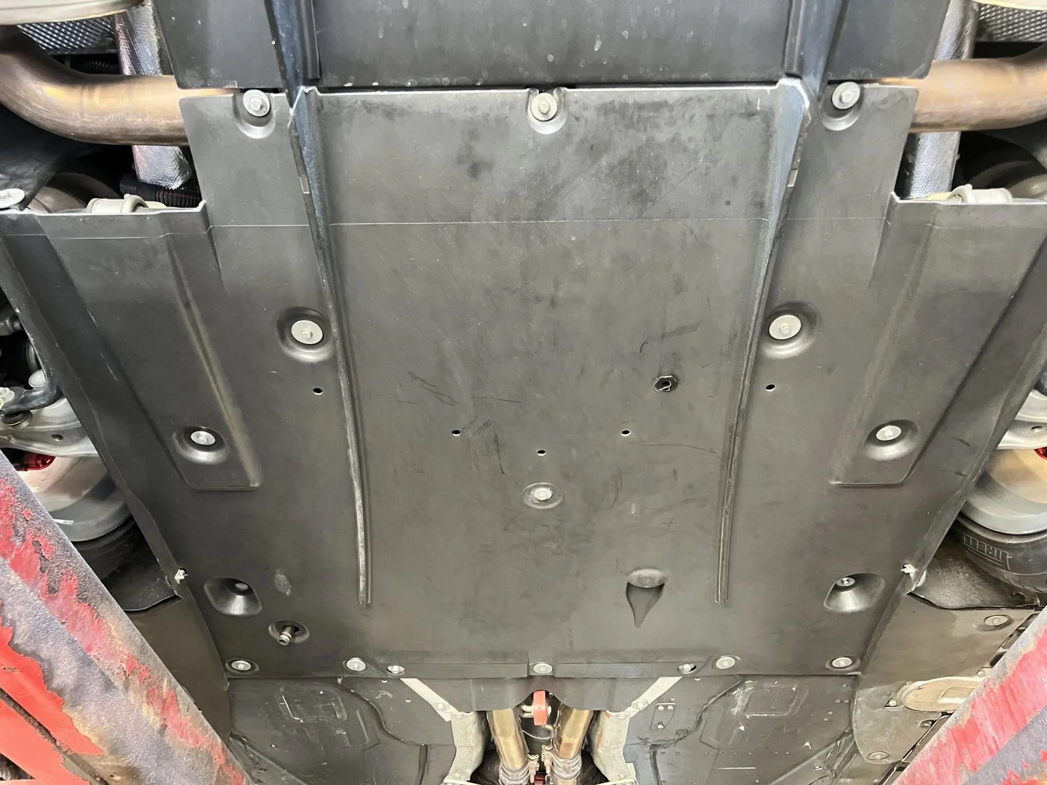 Underside of a vehicle showing a black metal skid plate with multiple bolts, part of the vehicle's undercarriage.
