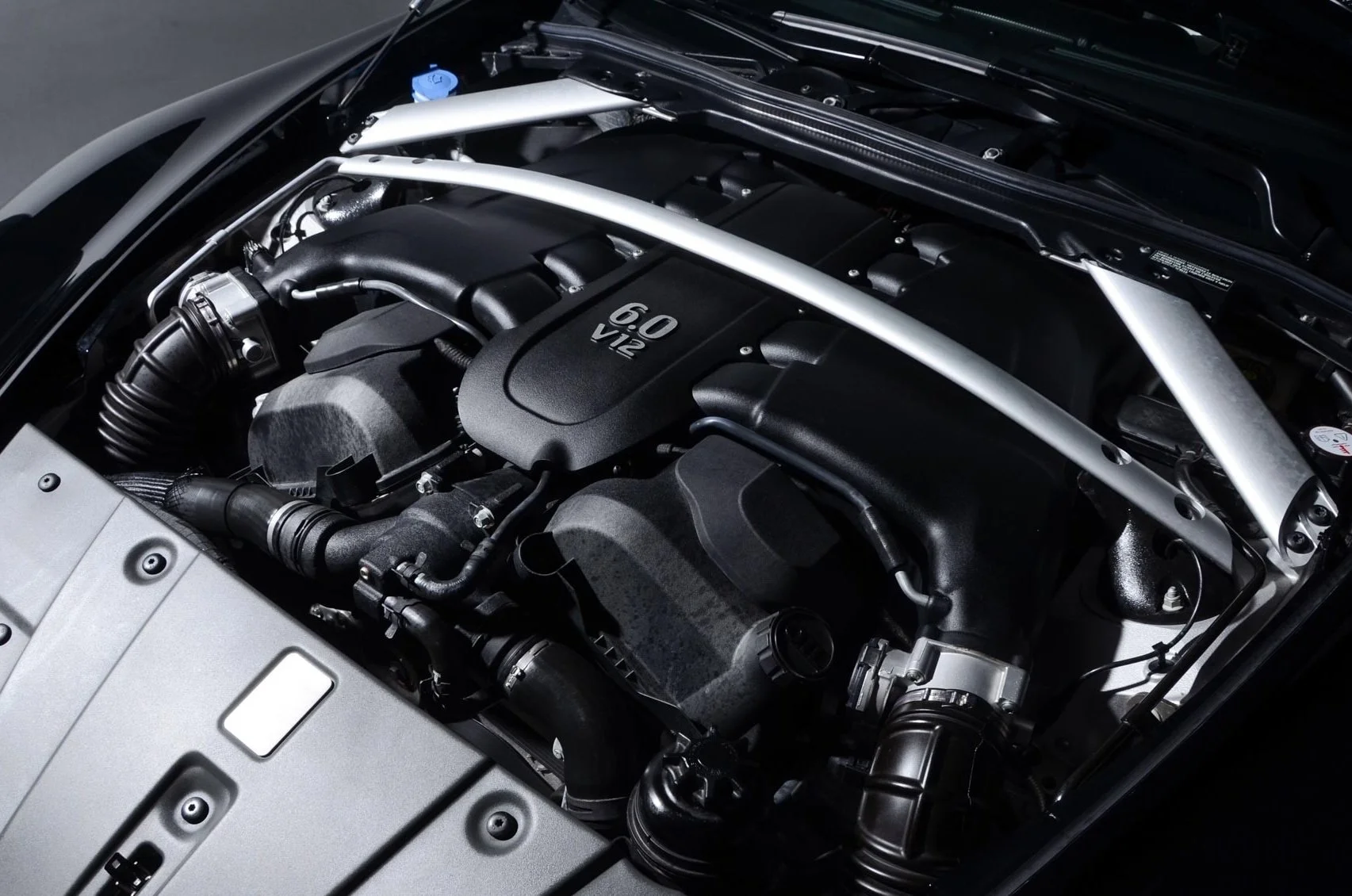 Close-up of a car engine with a black cover labeled '60 V12' and various hoses and metal components surrounding it.