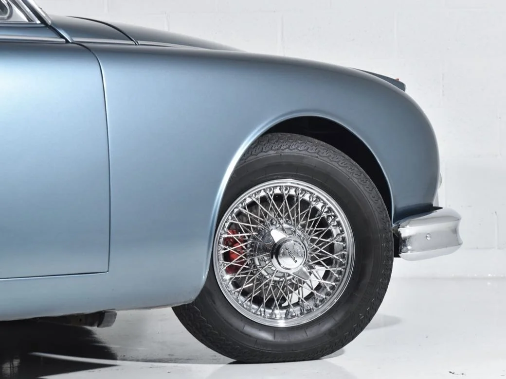 Close-up of the front wheel and part of the front fender of a vintage blue car with a wire-spoke wheel and a hubcap that reads 'JAGUAR.'