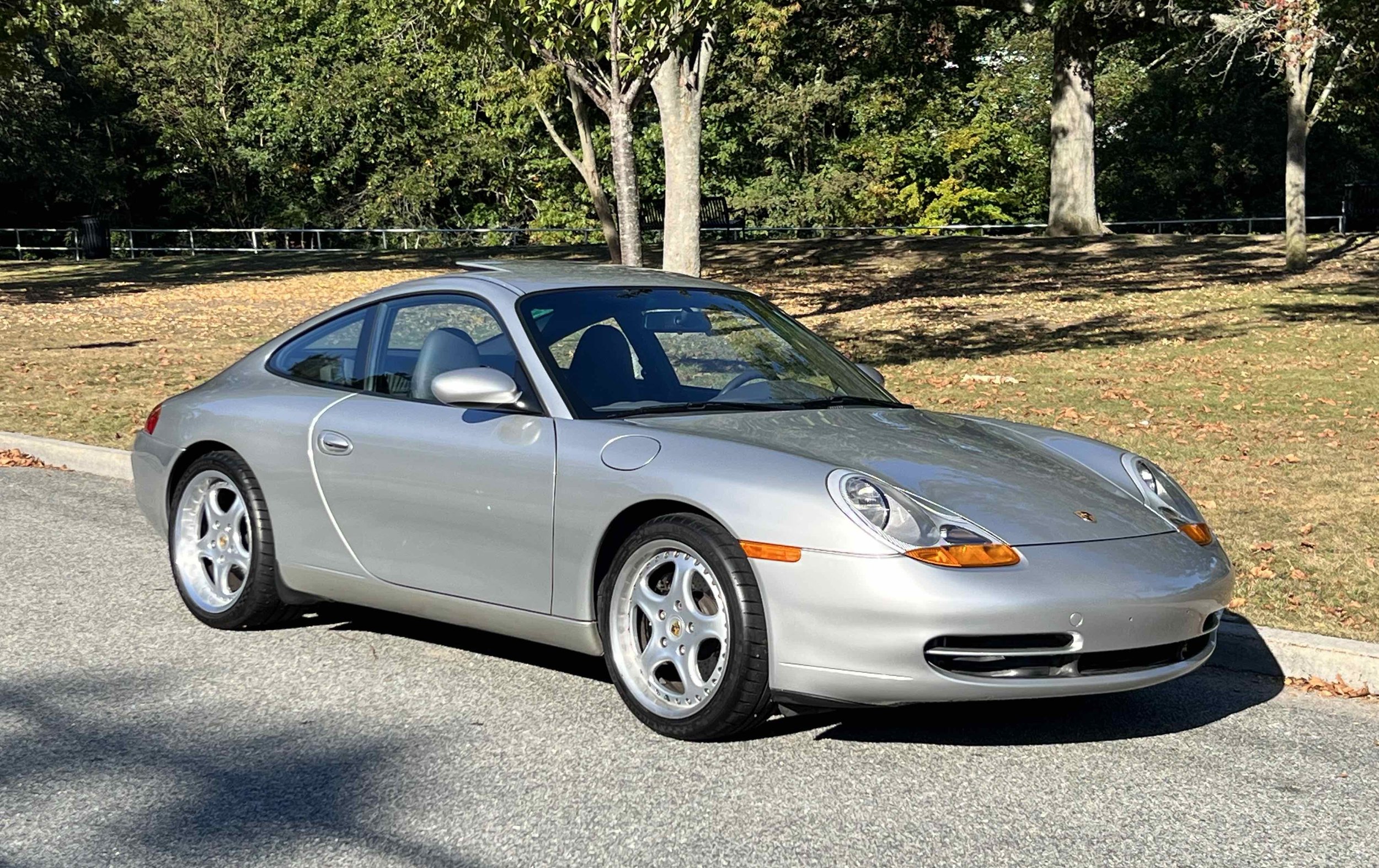 Silver Porsche sports car parked on the side of the road in a park with trees and fallen leaves in the background.